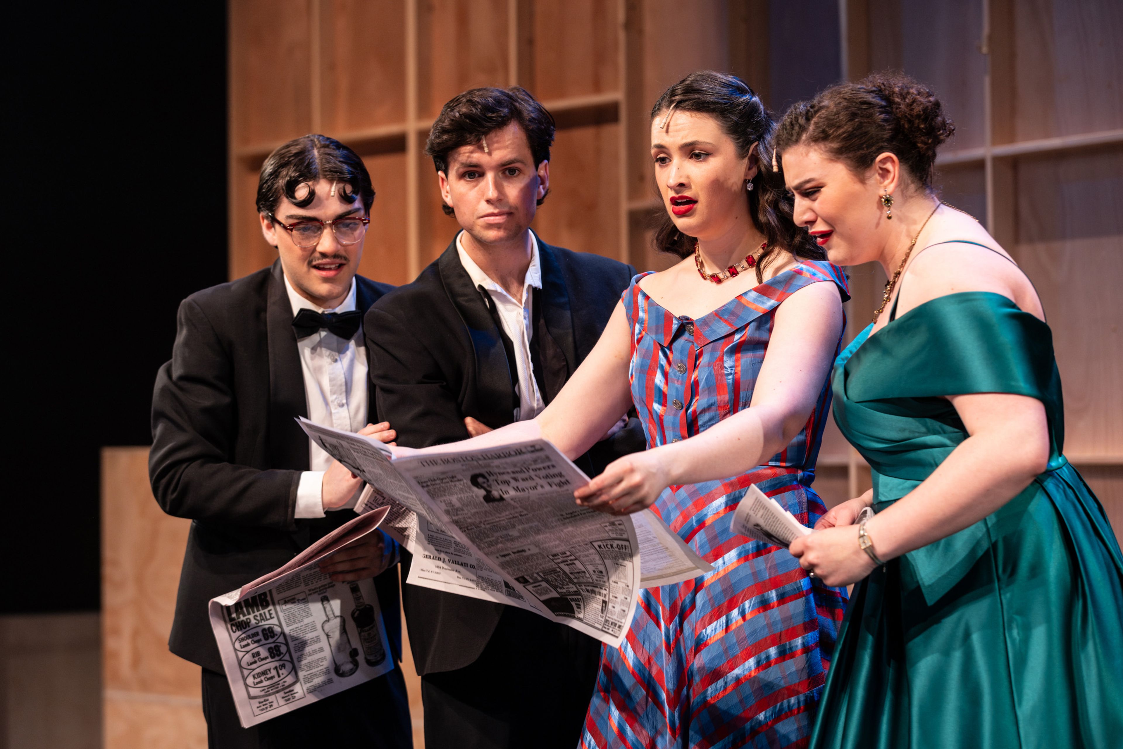 Theatre production photograph showing four performers reading a vintage-style newspaper, with two men in formal suits and two women in 1950s-style dresses against a wooden backdrop.