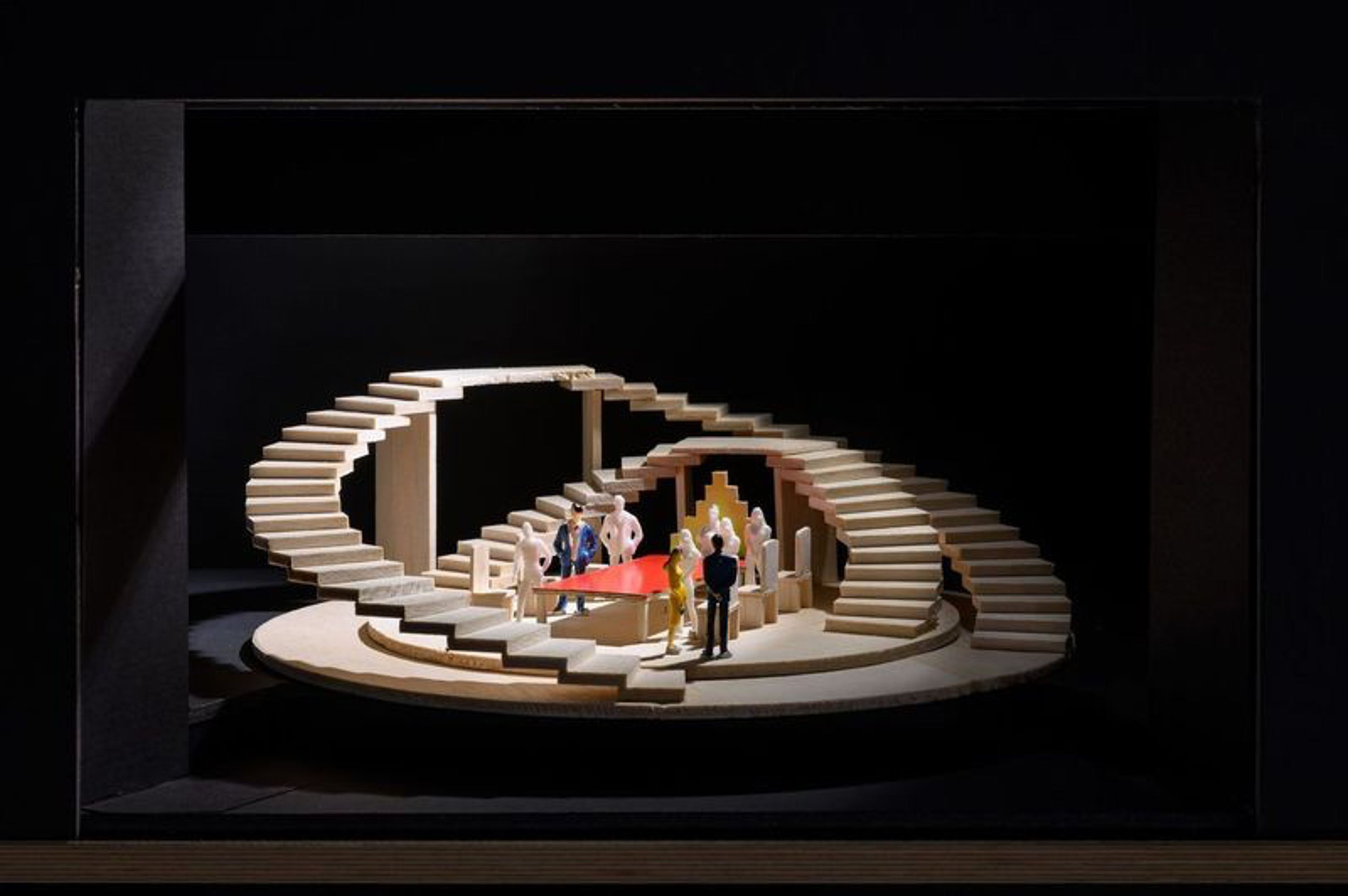 This is a stage model featuring a circular, multi-level platform with a grand, spiral staircase wrapping around the structure. At the center, a bright red triangular table is surrounded by small figurines