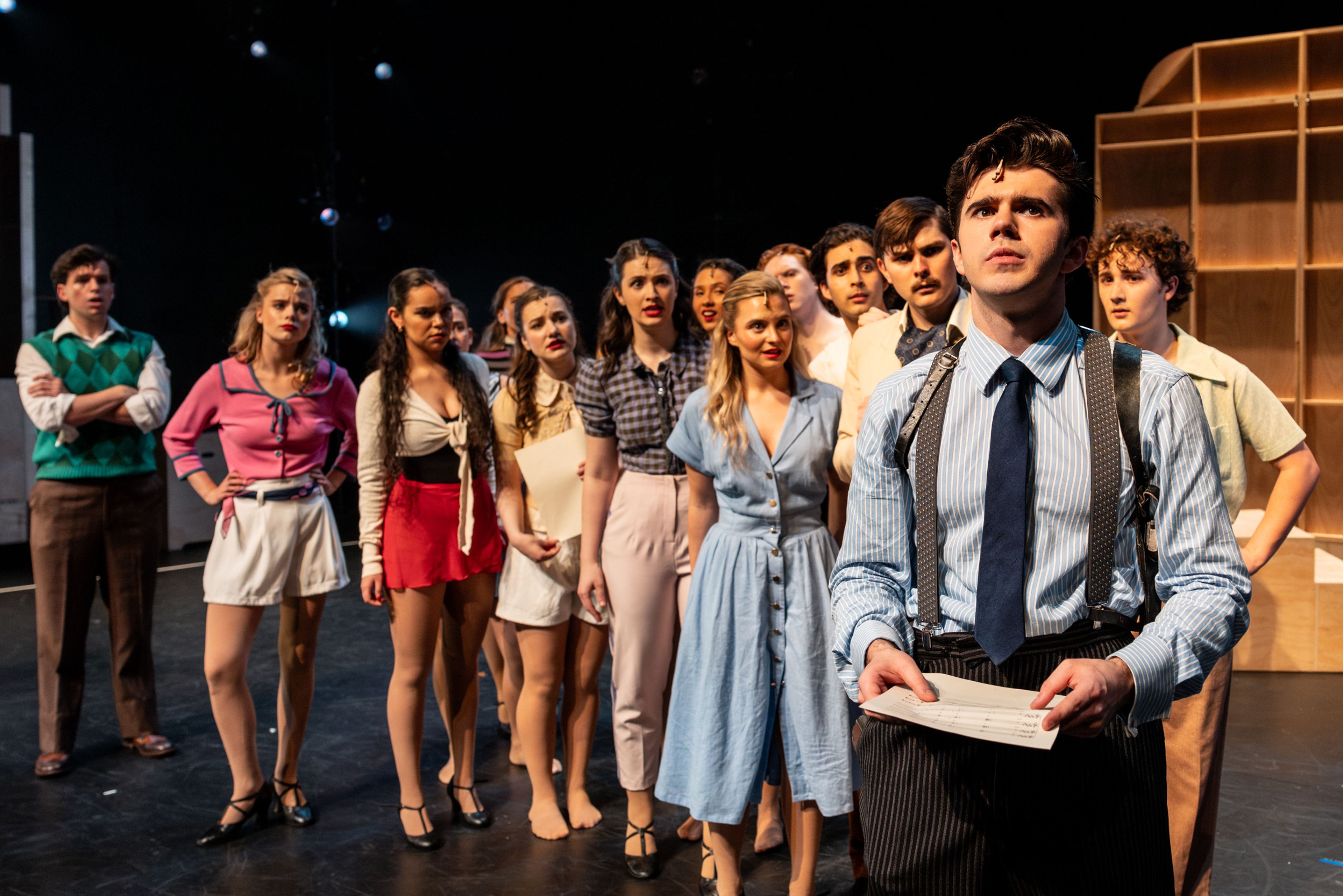 Theatre production photograph capturing a dramatic lineup of performers in 1950s daywear, with a young man in striped trousers and suspenders holding sheet music in the foreground.