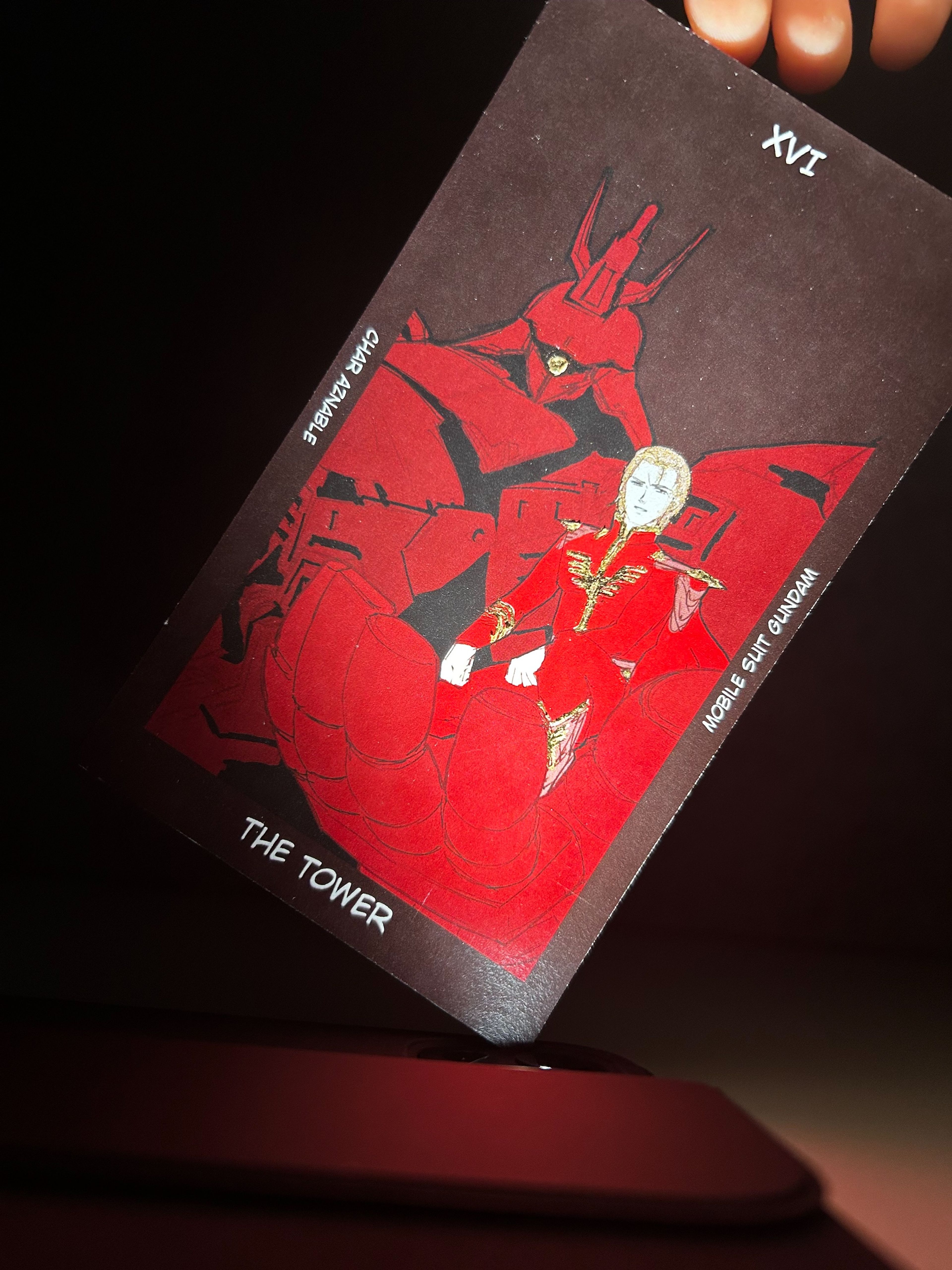 Close-up of a tarot card titled "The Tower" illustrated with a red-armoured figure and a character in regal red attire, titled "Char Aznable" from Mobile Suit Gundam.