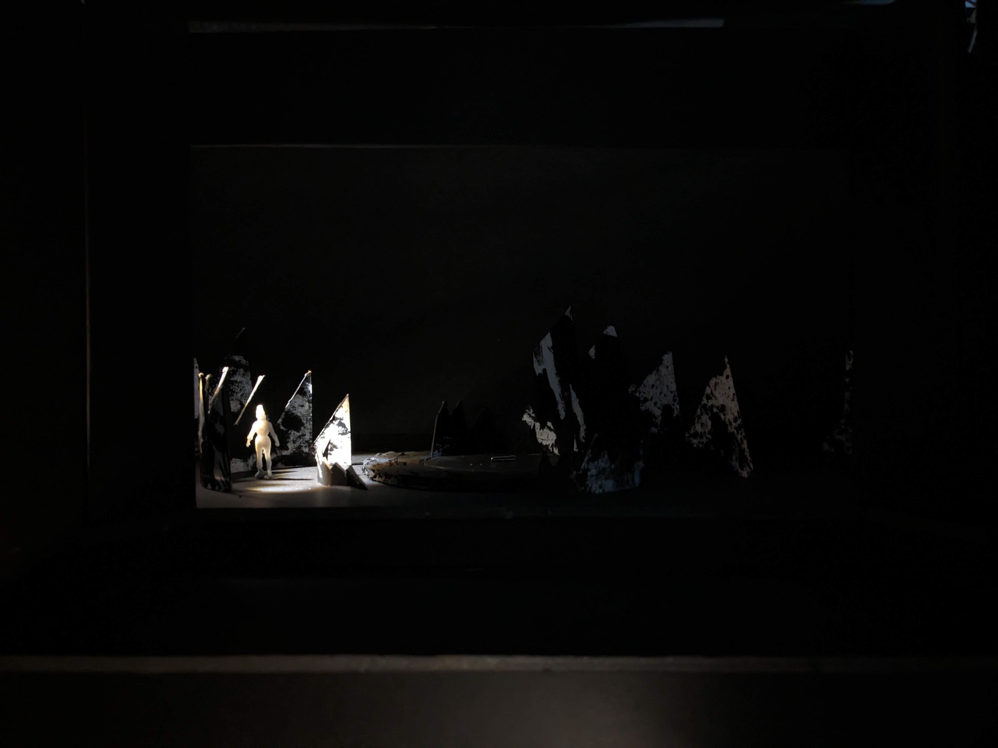 A solitary white figure illuminated on the left side of a dark set model with jagged mountain cutouts.