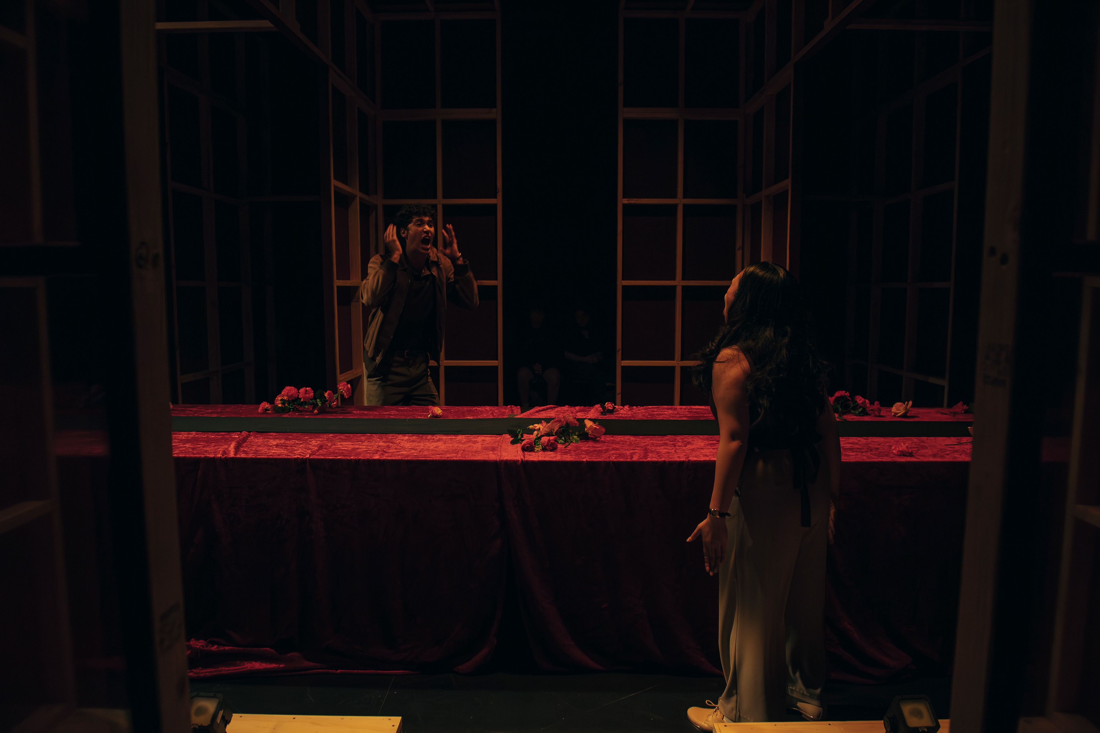 Two performers engage in an intense dramatic scene inside a dimly lit, structured stage setup. A long, red velvet-covered table adorned with scattered roses separates them