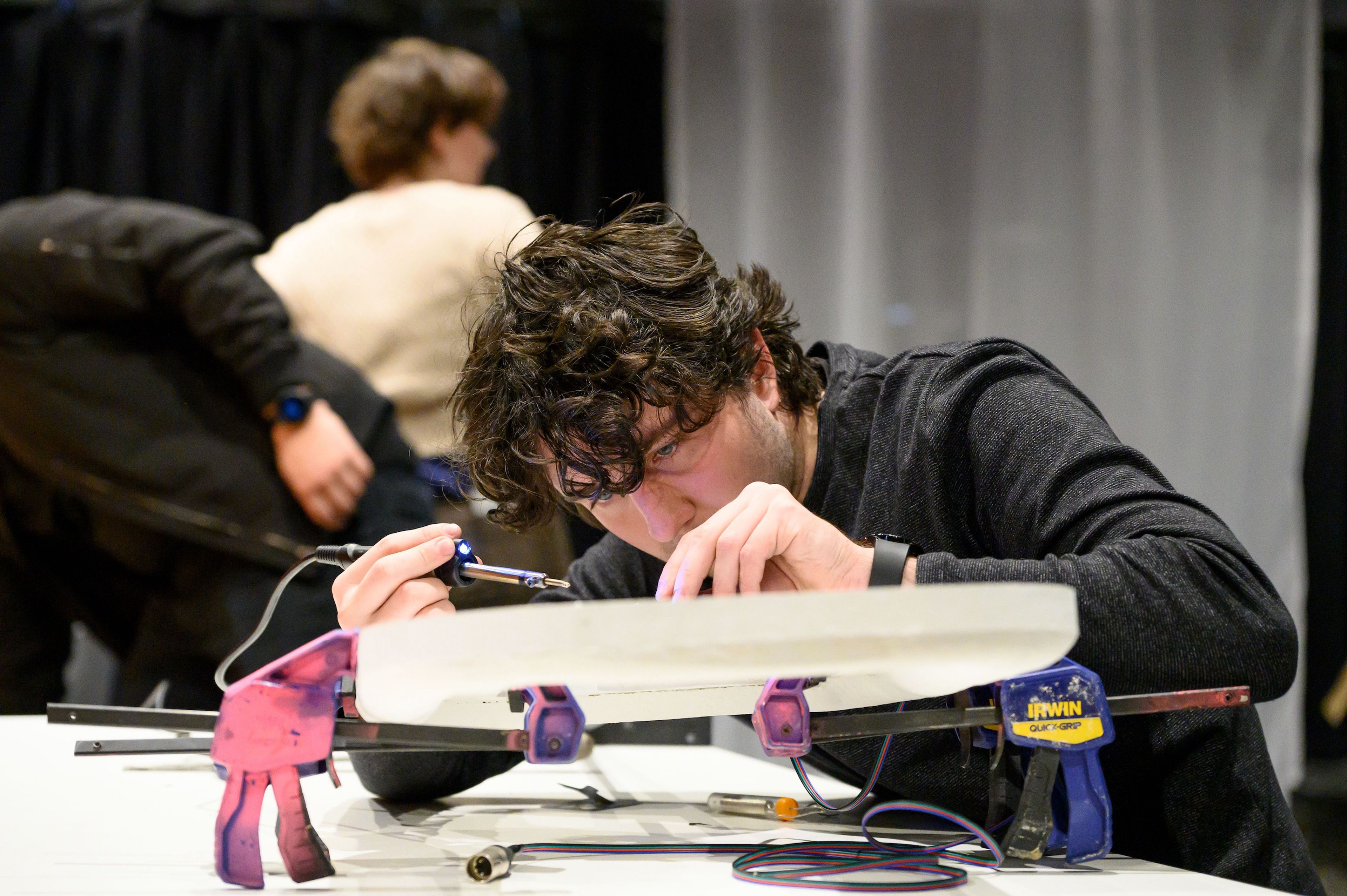 A person intently works on a technical project, holding a small tool over a clamped piece of equipment on a worktable.