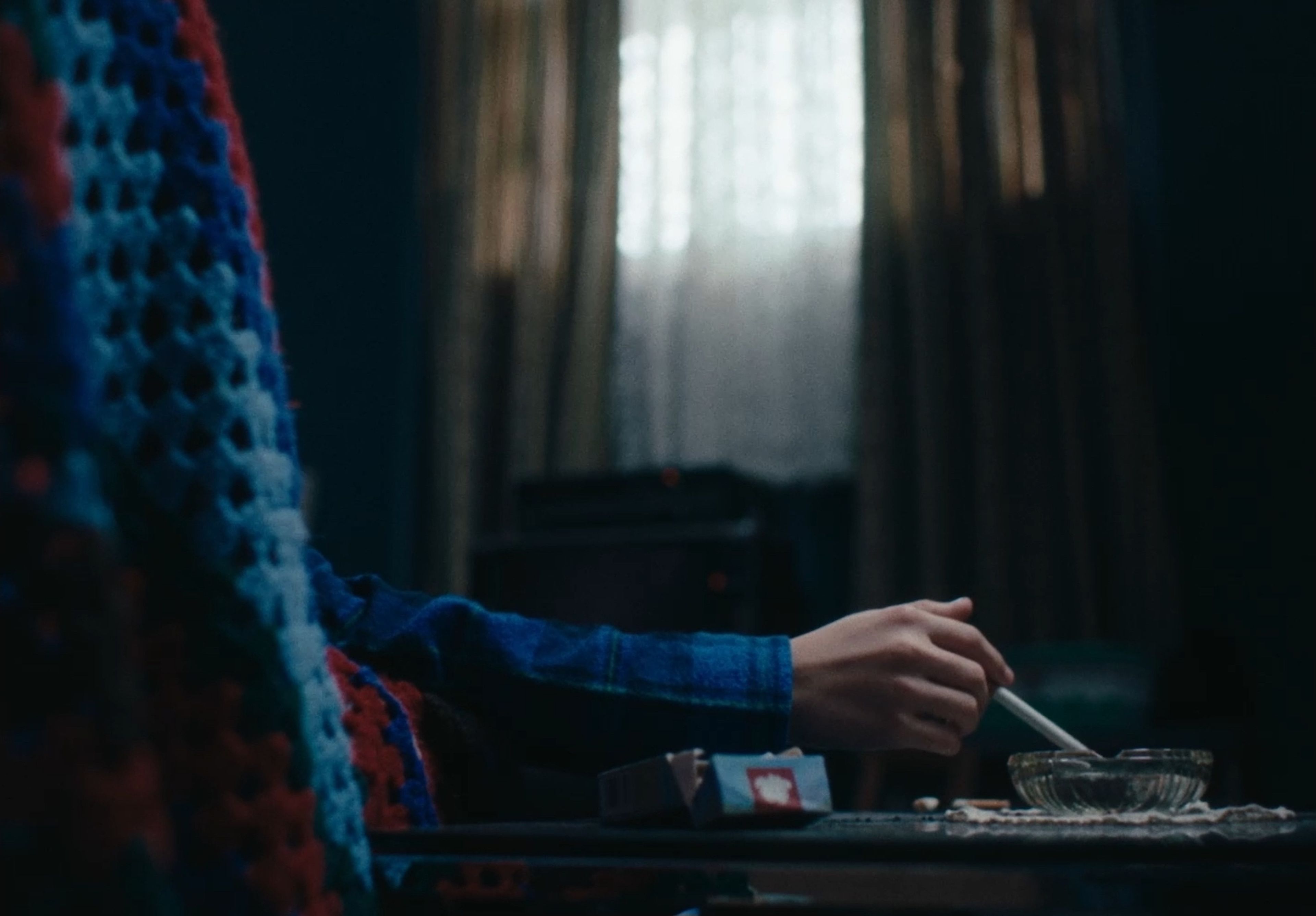 Film still showing a close-up of a hand in a blue and red patterned jumper holding a cigarette, with soft window light in the background.