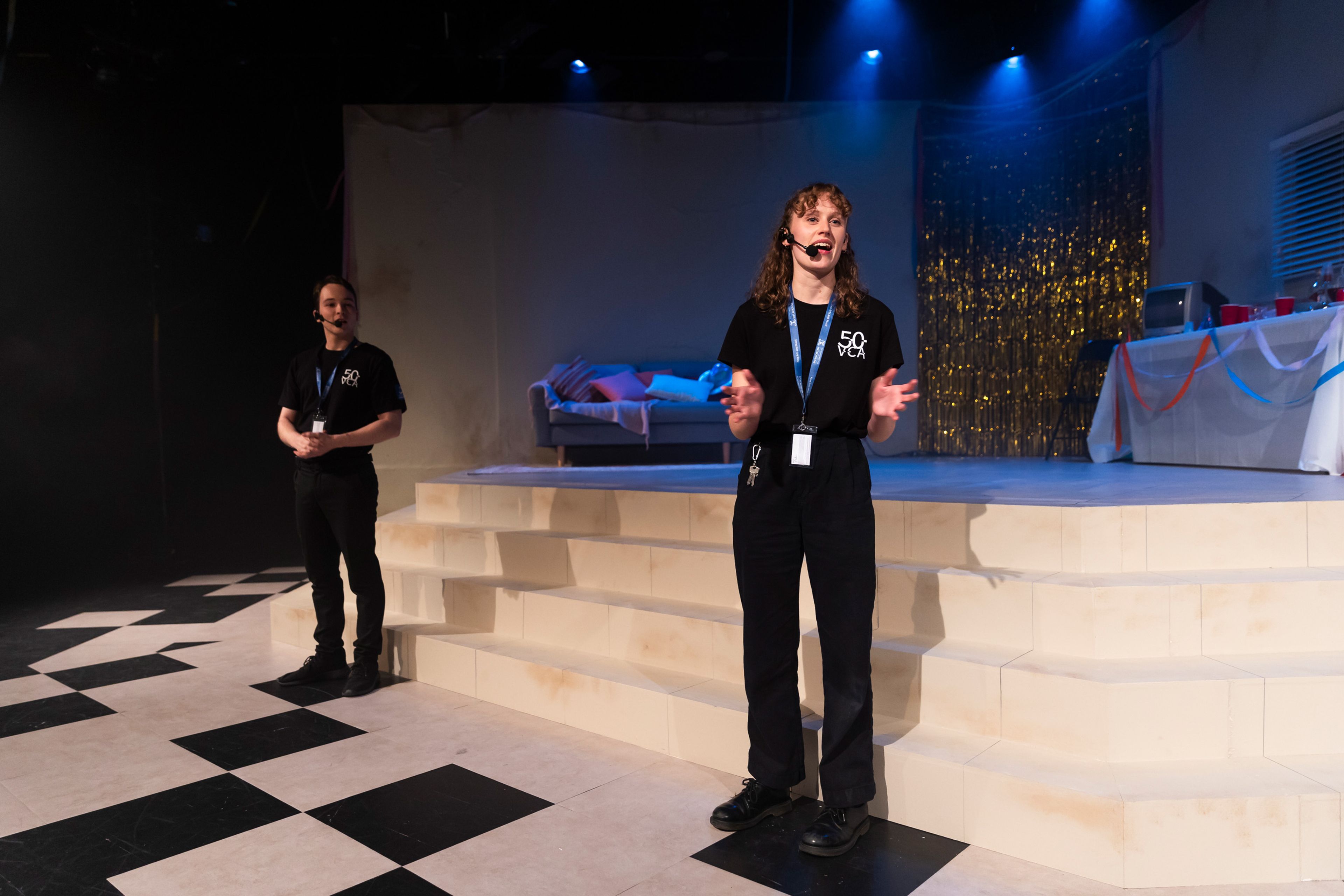 Theatre production photograph featuring two stage managers or crew members in black '50 VCA' t-shirts speaking to the audience, with a party setting visible behind them including gold tinsel and a couch.