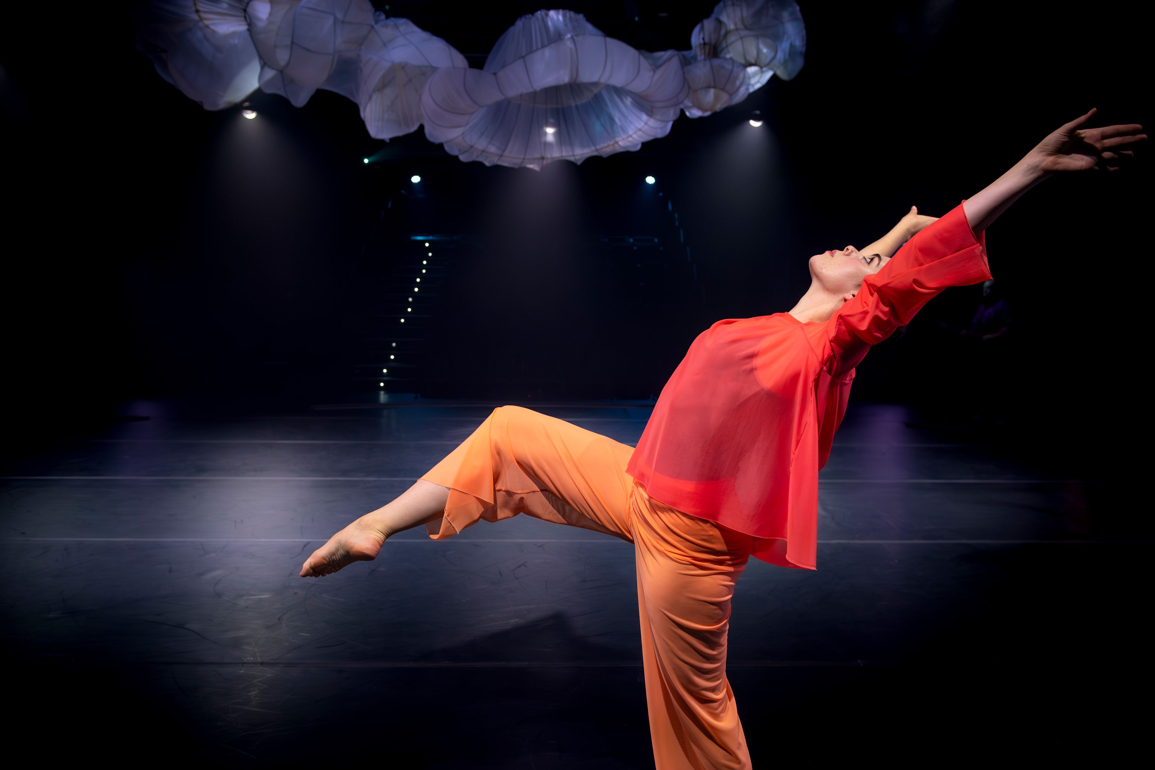 Theatre production photograph featuring a single dancer in vibrant orange and red costume stretching gracefully with arms extended, under sculptural fabric forms and surrounded by moody lighting.