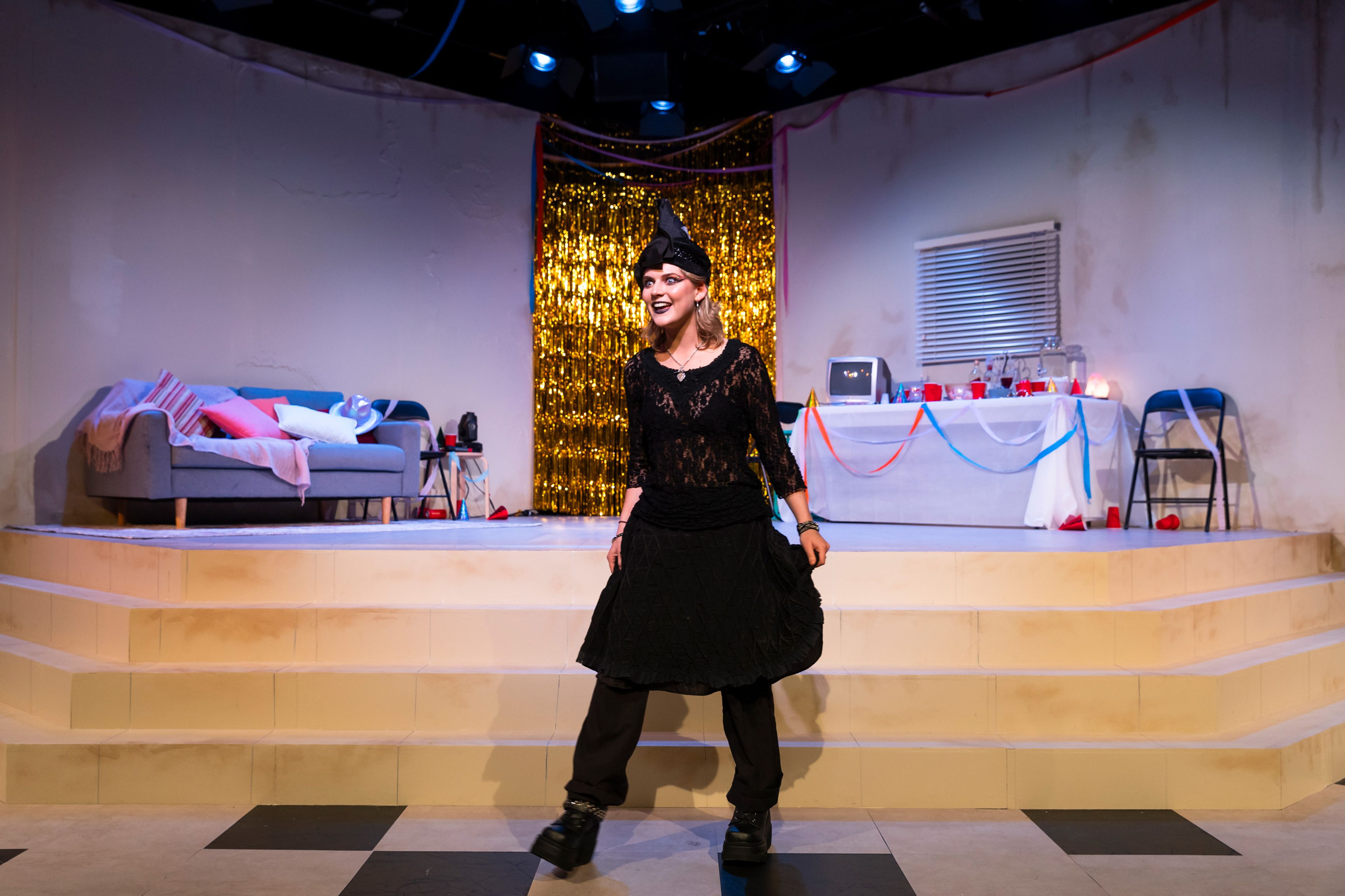 Theatre production photograph showing a performer in a black party outfit and witch's hat on a checkerboard floor, with a gold tinsel backdrop and party decorations visible.