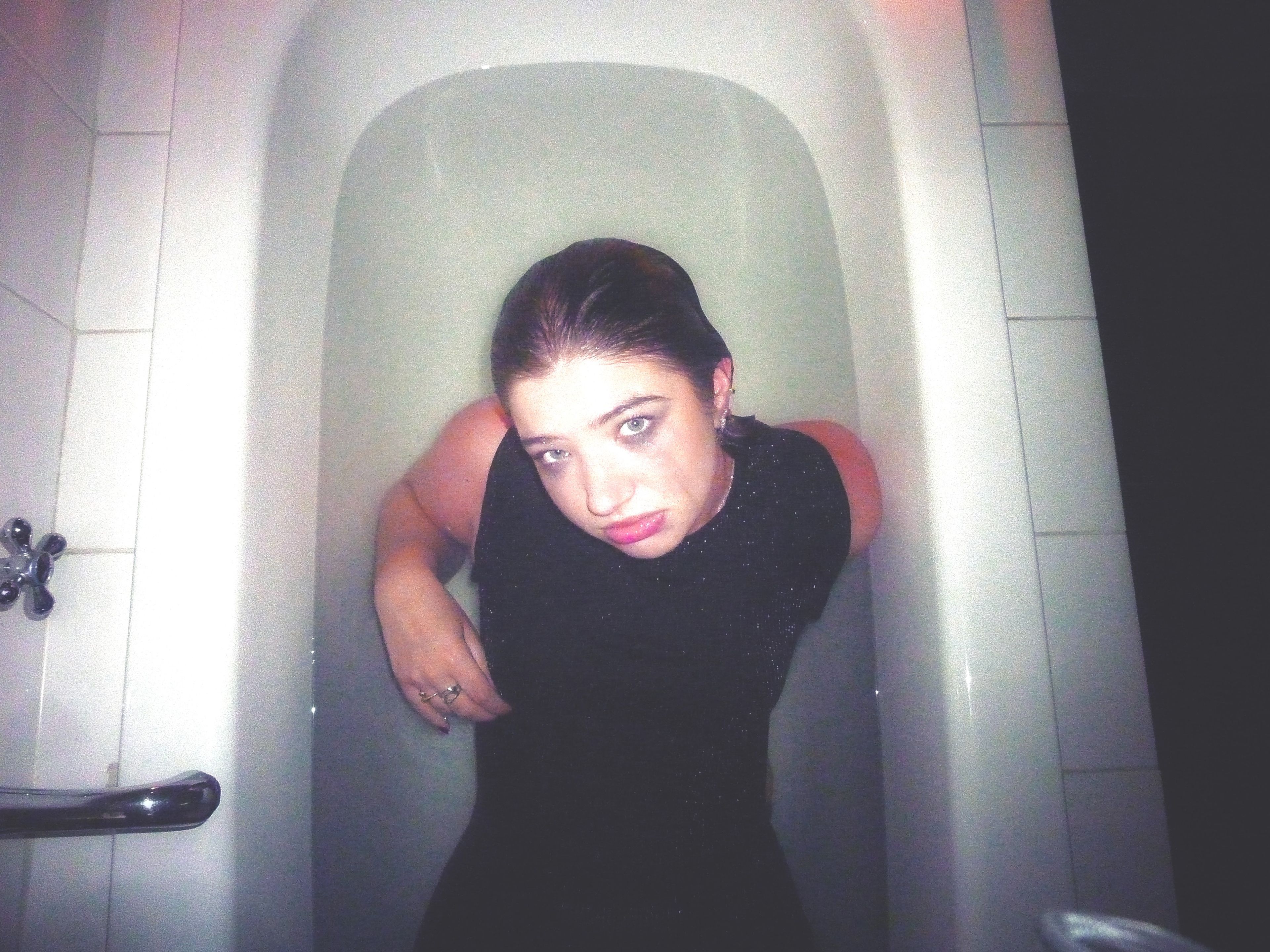 The same woman is now captured fully clothed in a bathtub, with her arms partially submerged. Her expression is contemplative, and the stark lighting contrasts with the white bathroom tiles, evoking a feeling of solitude and introspection.