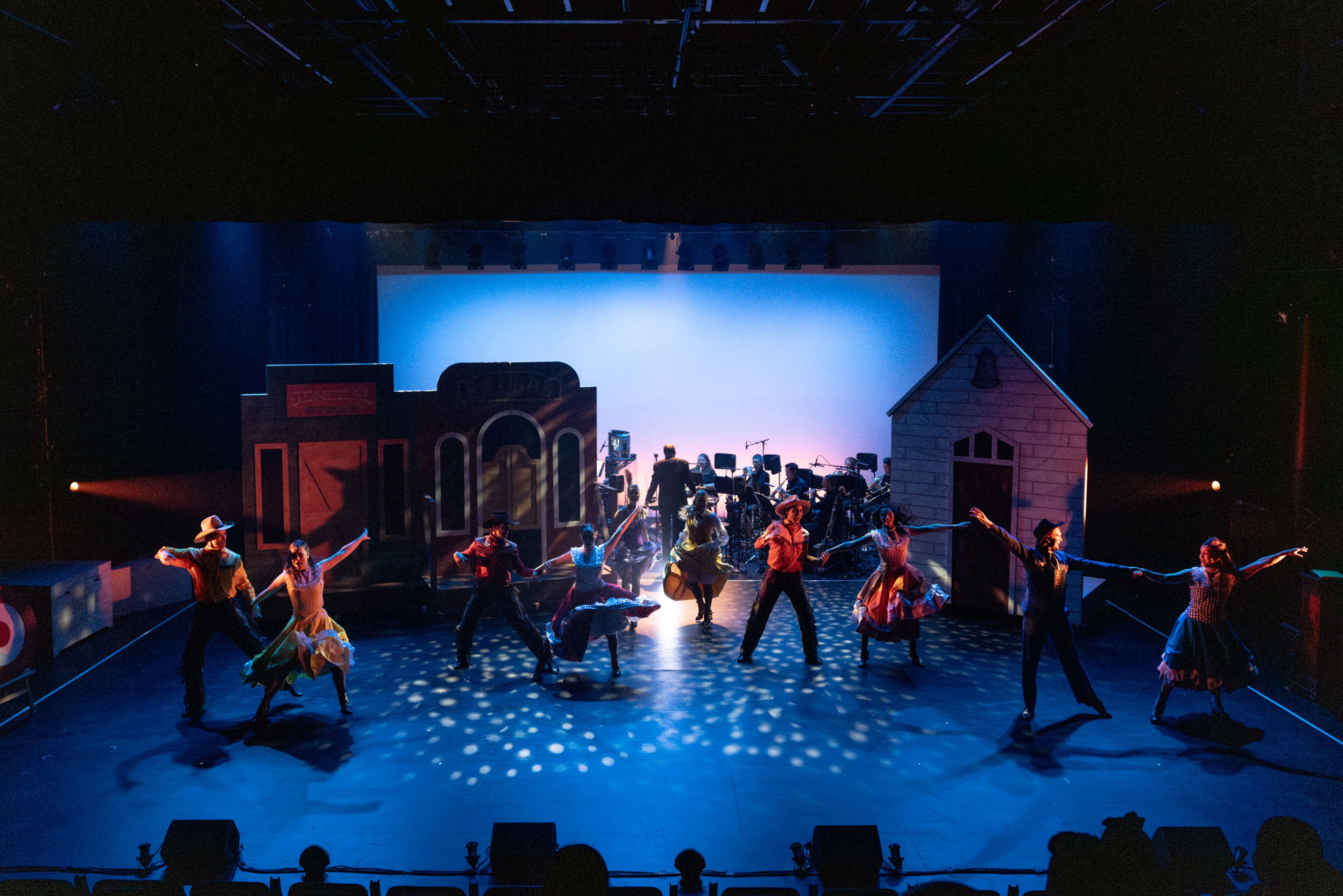 Theatre production photograph showing a Western-themed dance number with performers in cowboy costumes and colourful skirts, performing in front of a saloon set with stage lighting creating spotted patterns on the floor.