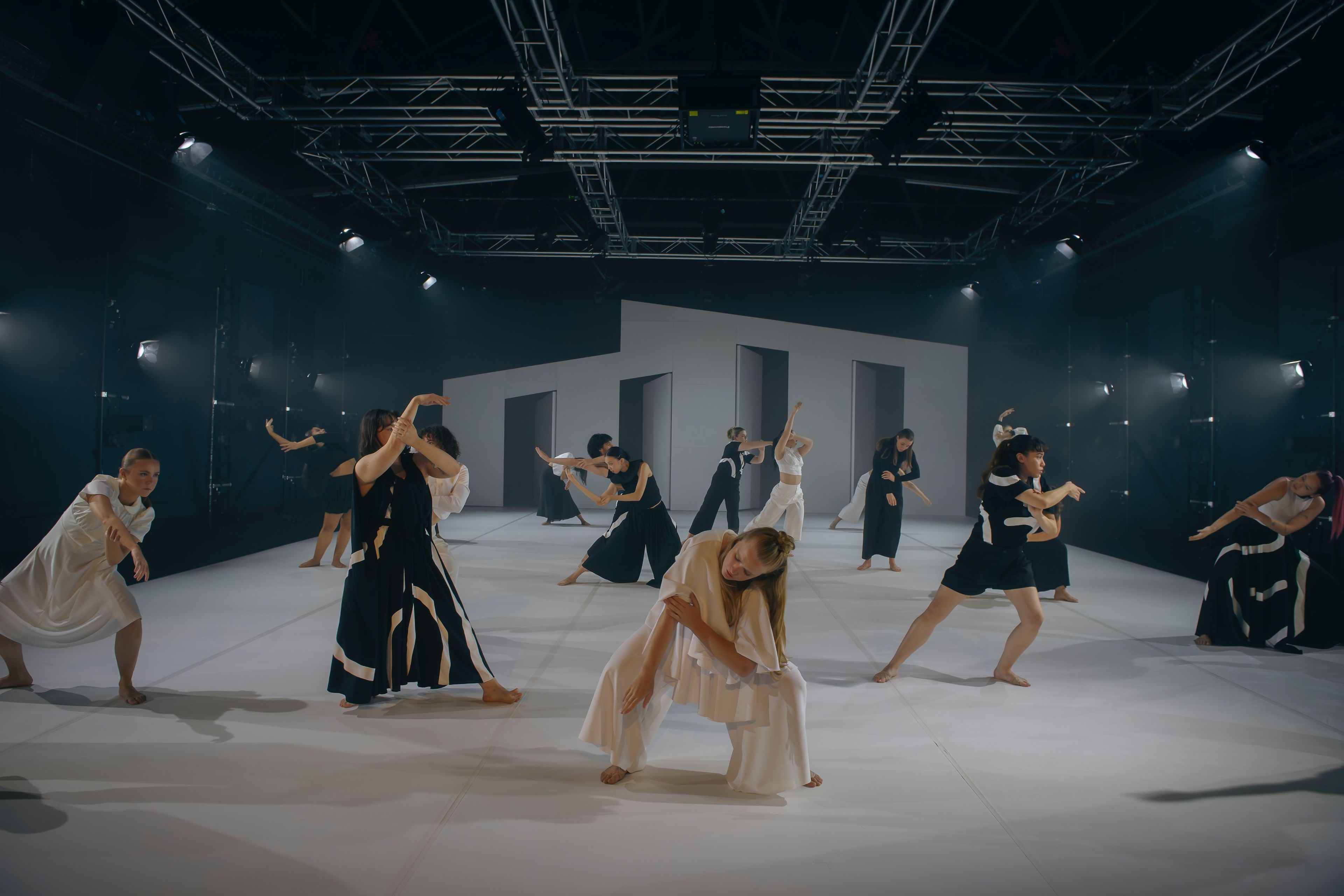 Theatre production photograph showing a group of dancers in black and white costumes, performing expressive movements on a white stage with industrial lighting overhead.