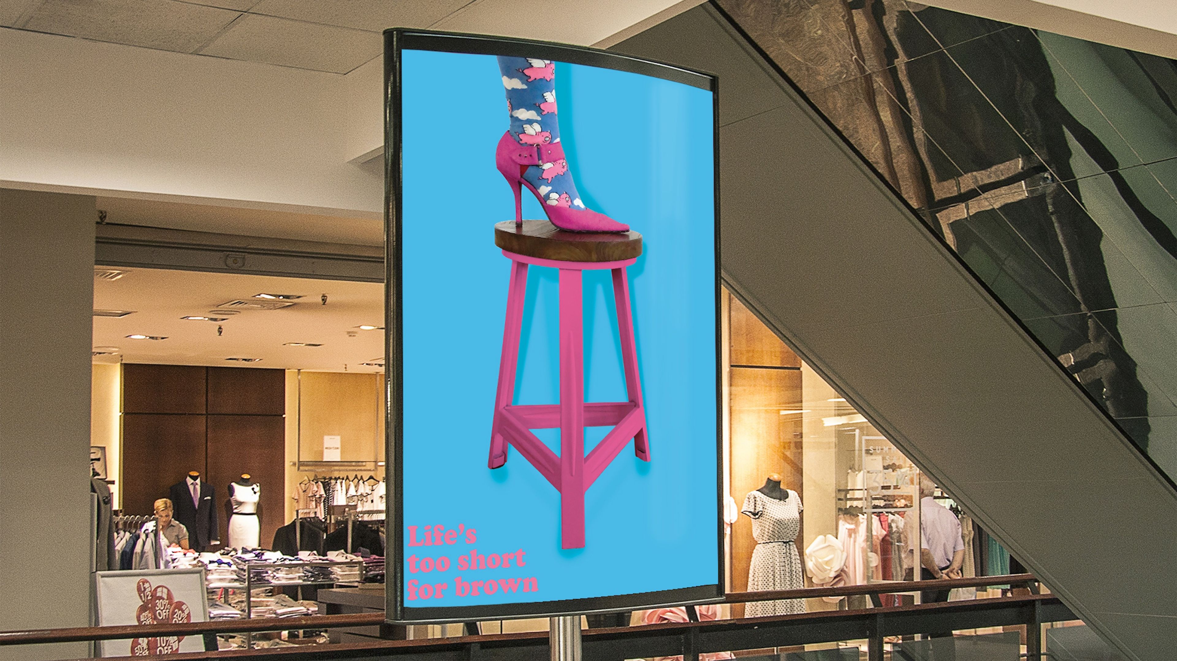 Digital display advertisement in a shopping centre, showing a bright pink high heel on a matching pink stool against a vivid blue background. The text in pink reads, 'Life’s too short for brown,' adding a playful, fashion-forward message.