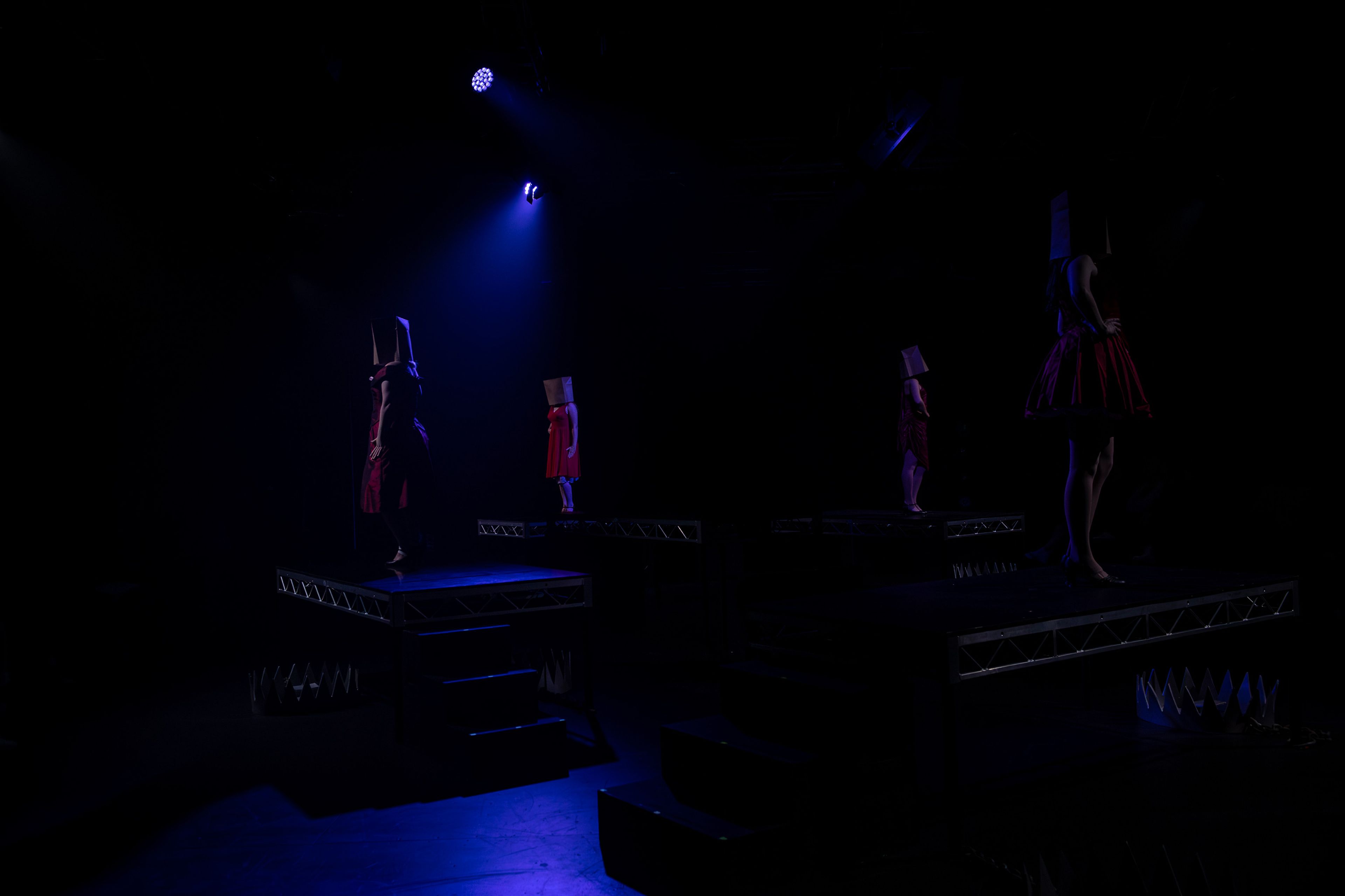 Performers wearing paper bag masks and red dresses stand individually on elevated platforms under dim blue stage lighting