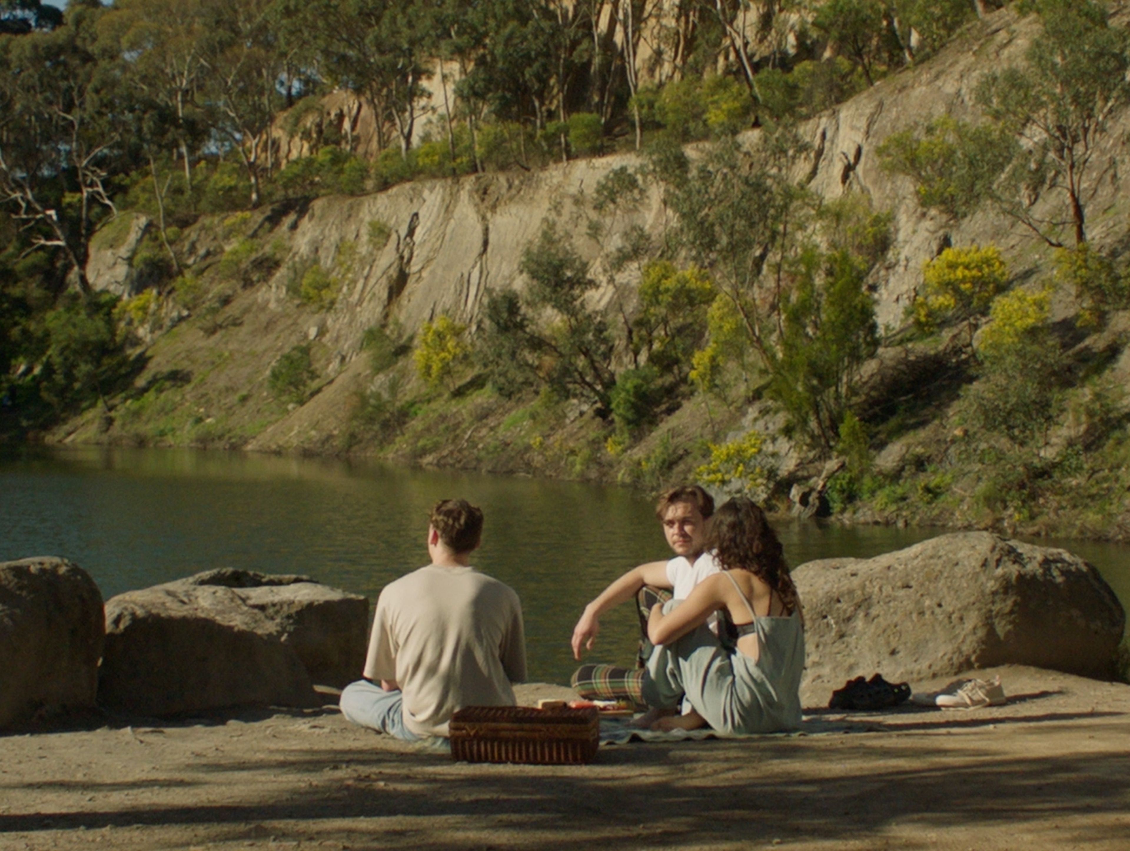 Film still showing three people having a picnic by a river, with eucalyptus trees and rocky cliffs in the background, shot in warm afternoon light.