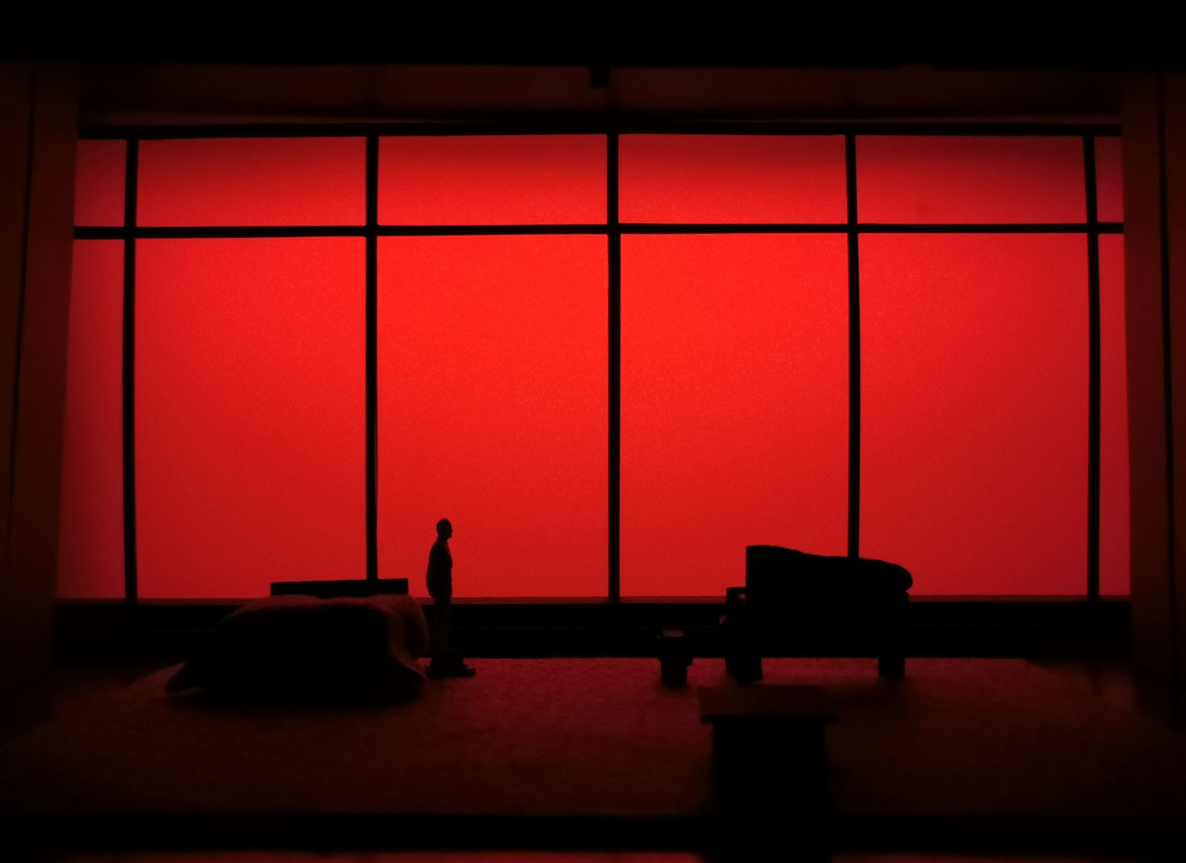 Scale model of a set design featuring a figure standing on a dark stage with a vibrant red backdrop.