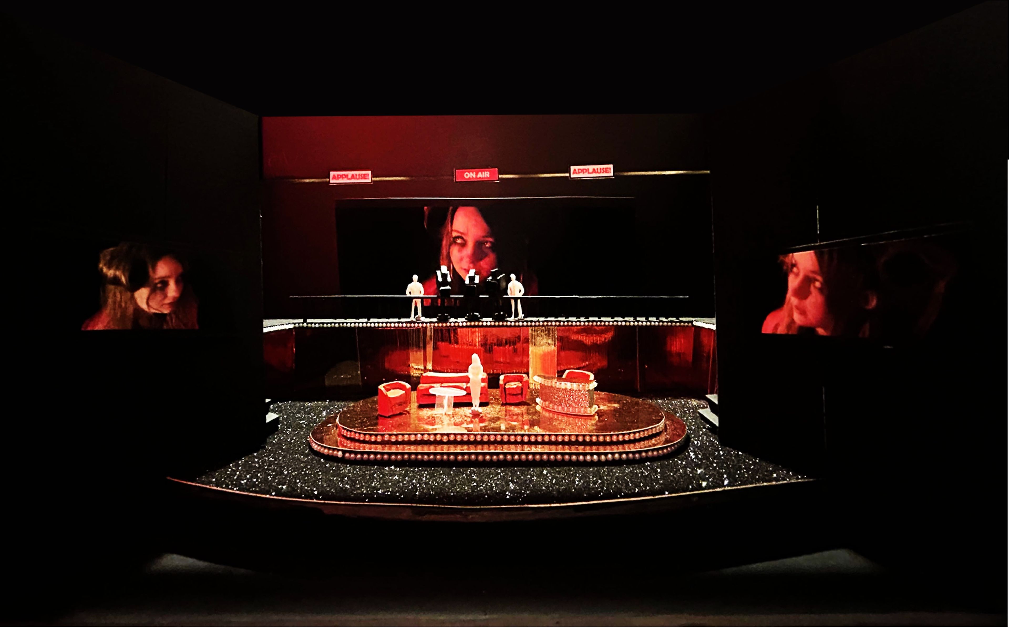 Set model featuring a red and black stage with 'applause' and 'on air' signs, multiple screens displaying close-ups of a woman's face, and miniature figures on stage.