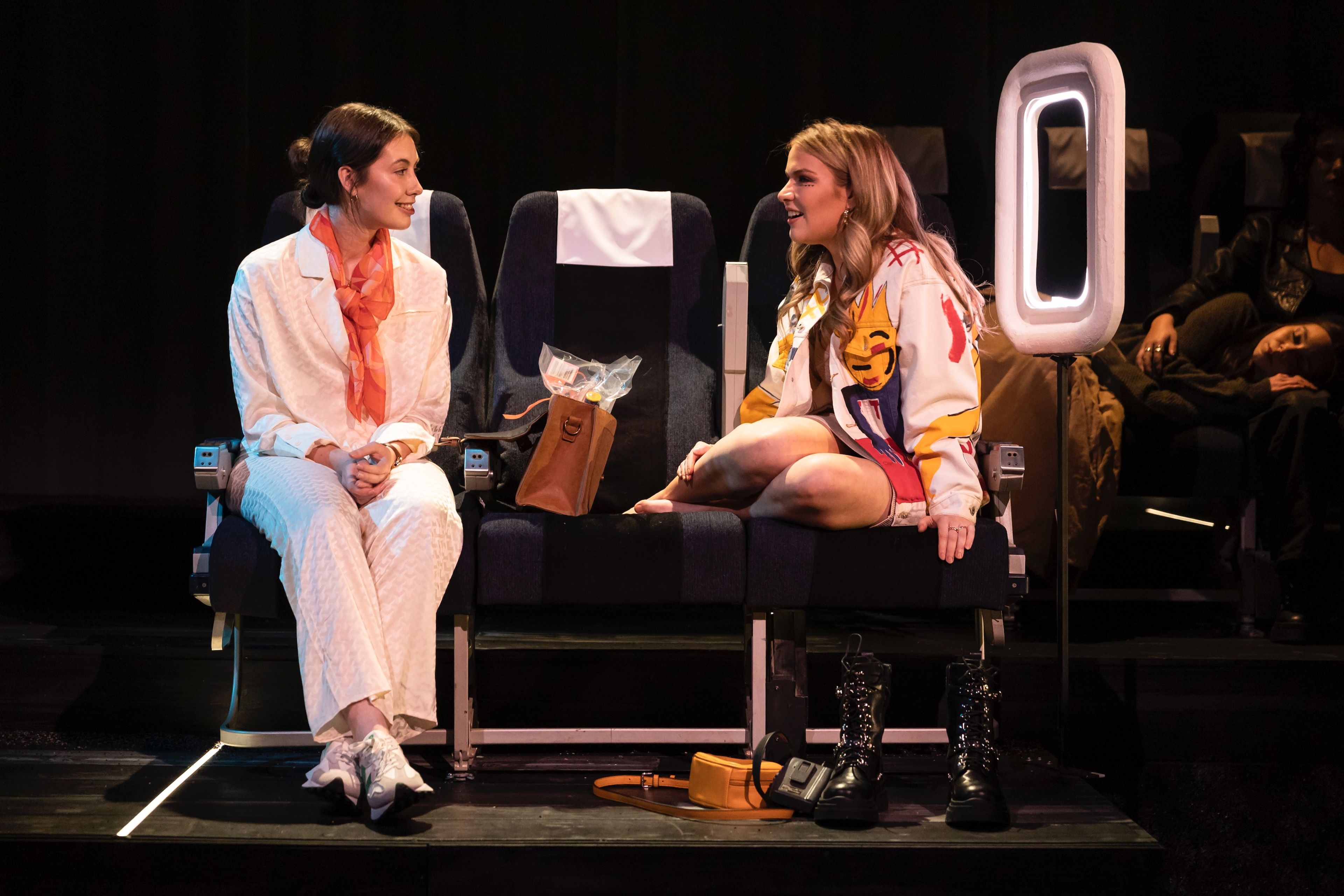 Two actors in a set designed to resemble an airplane cabin, sitting in adjacent seats, engaged in conversation, with a lit window frame beside them.