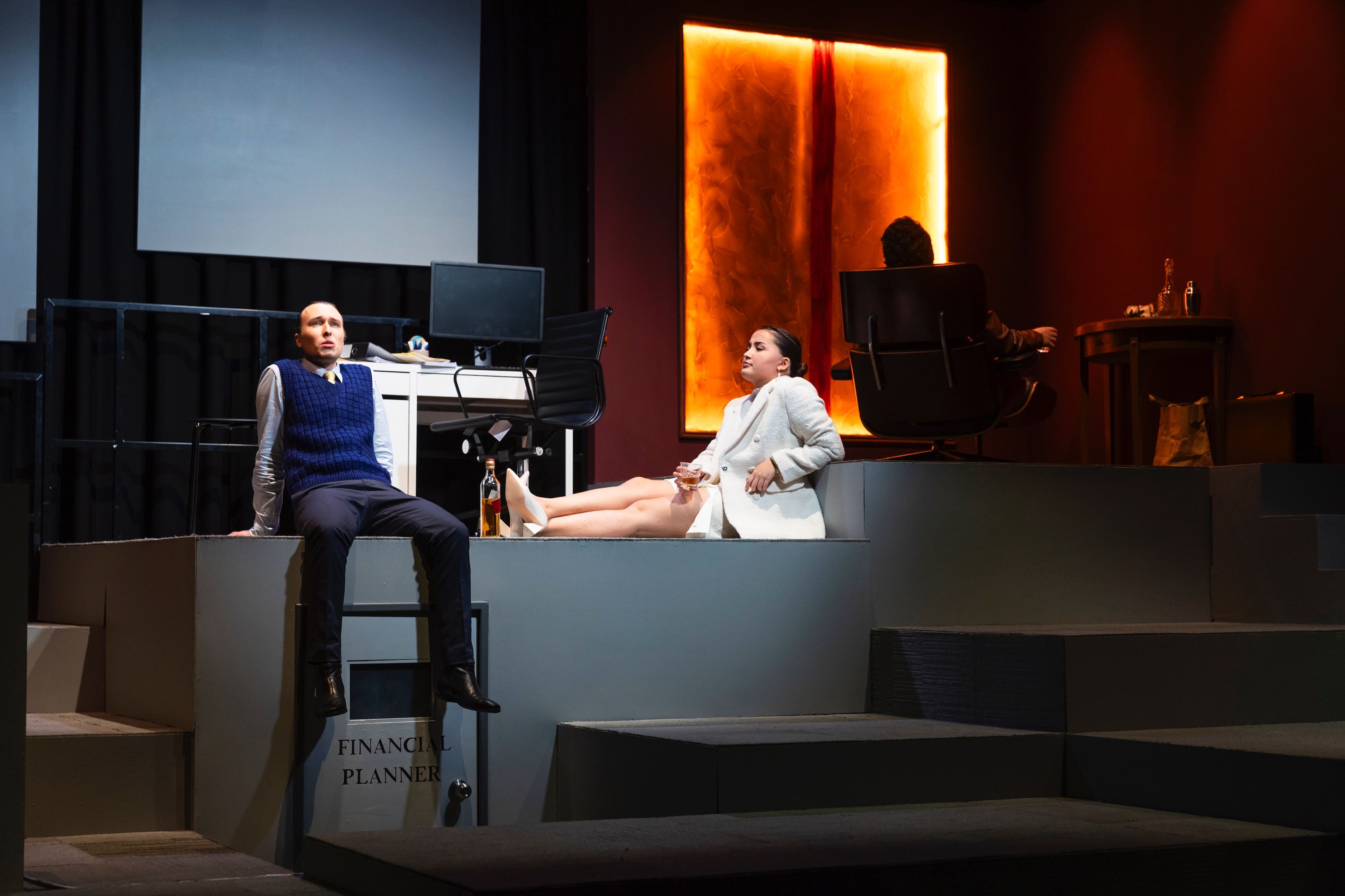 Theatre scene with a corporate office setup, where one actor in a white suit lounges with a drink while another sits beside a computer, under warm, amber-toned lighting.