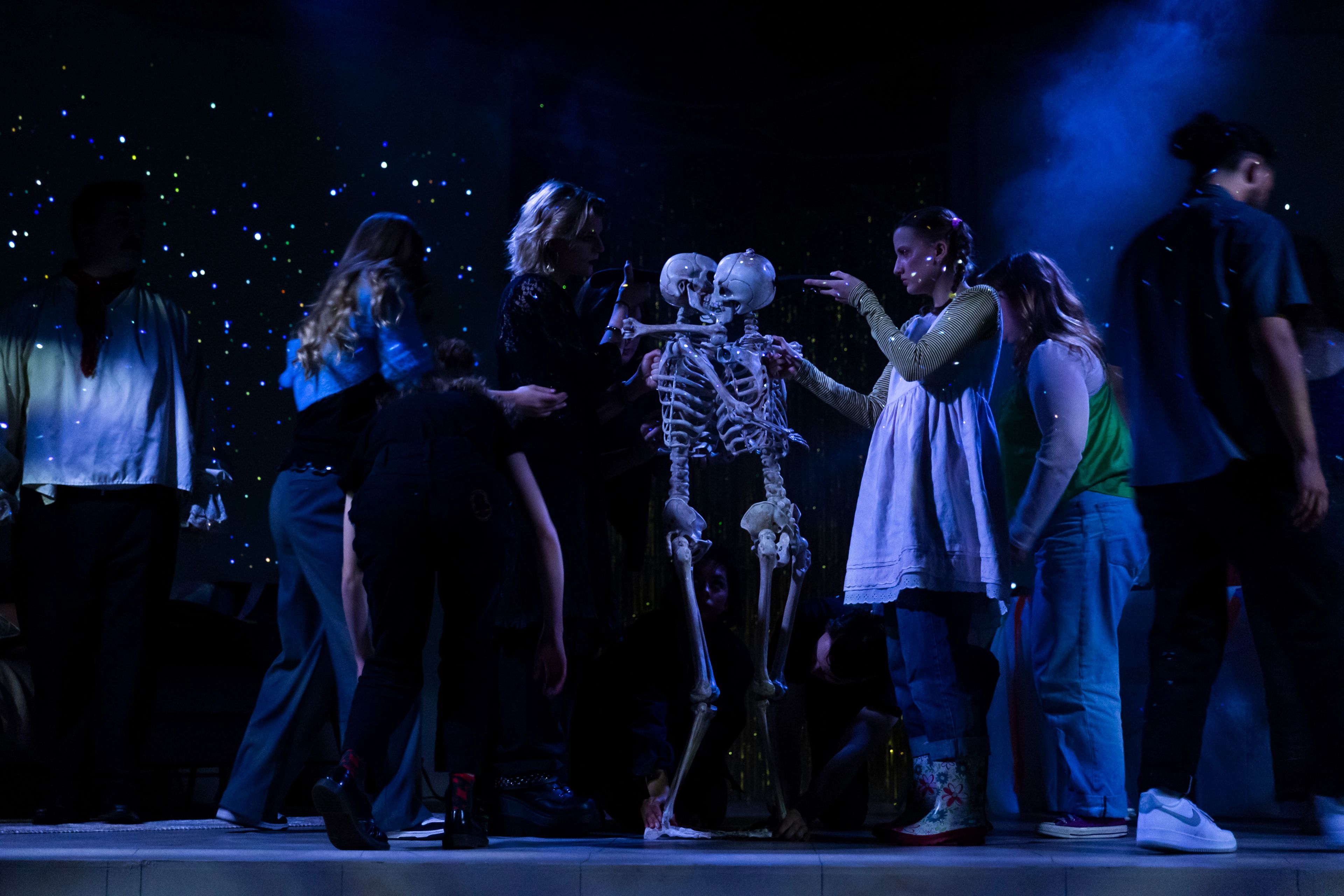 Theatre production photograph showing performers gathered around two anatomical skeleton props, lit dramatically in blue with star-like effects in the background.