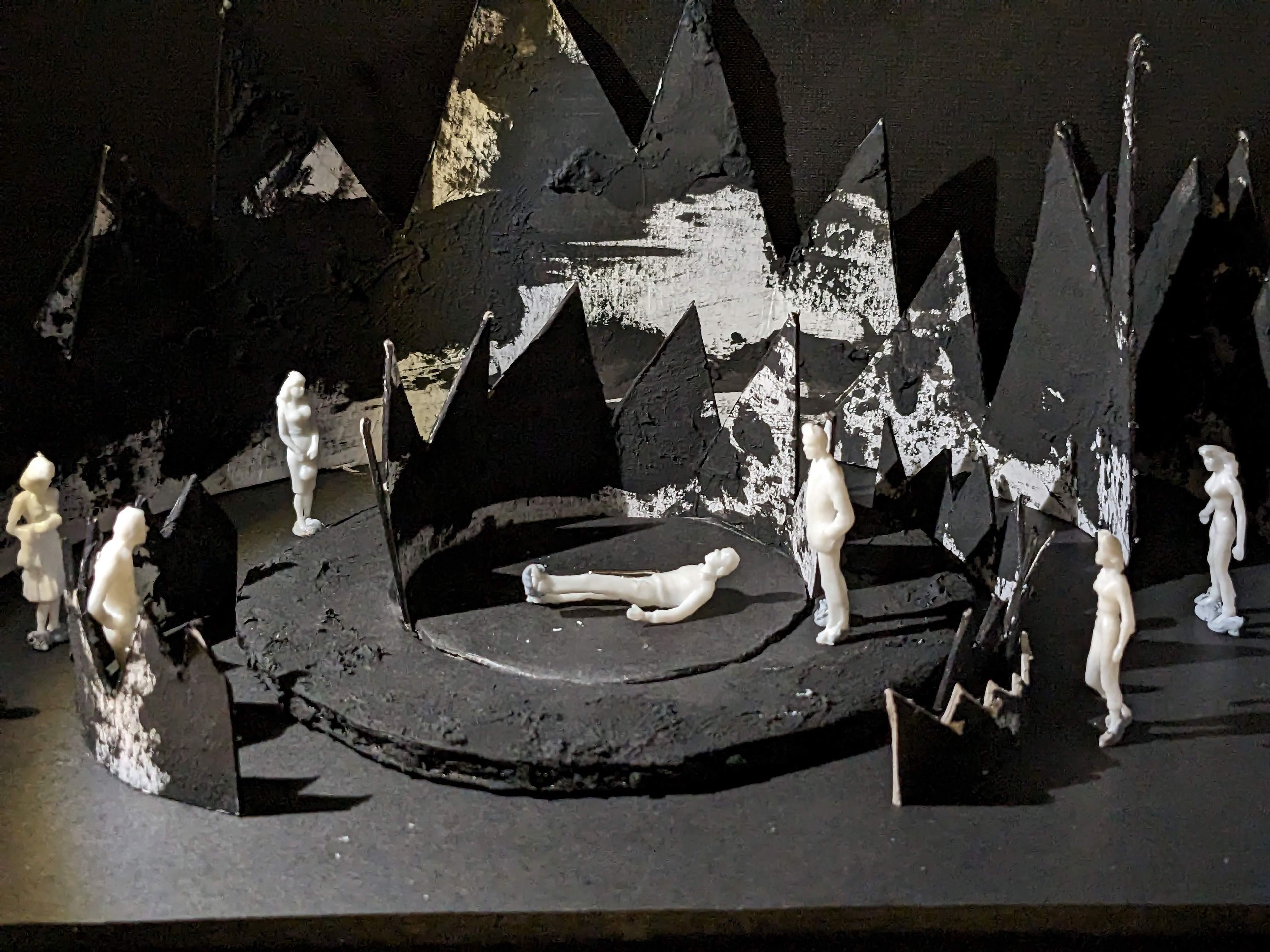 Set model showing white figures positioned on a rugged black circular stage, surrounded by abstract, pointed mountain shapes with a gritty texture.