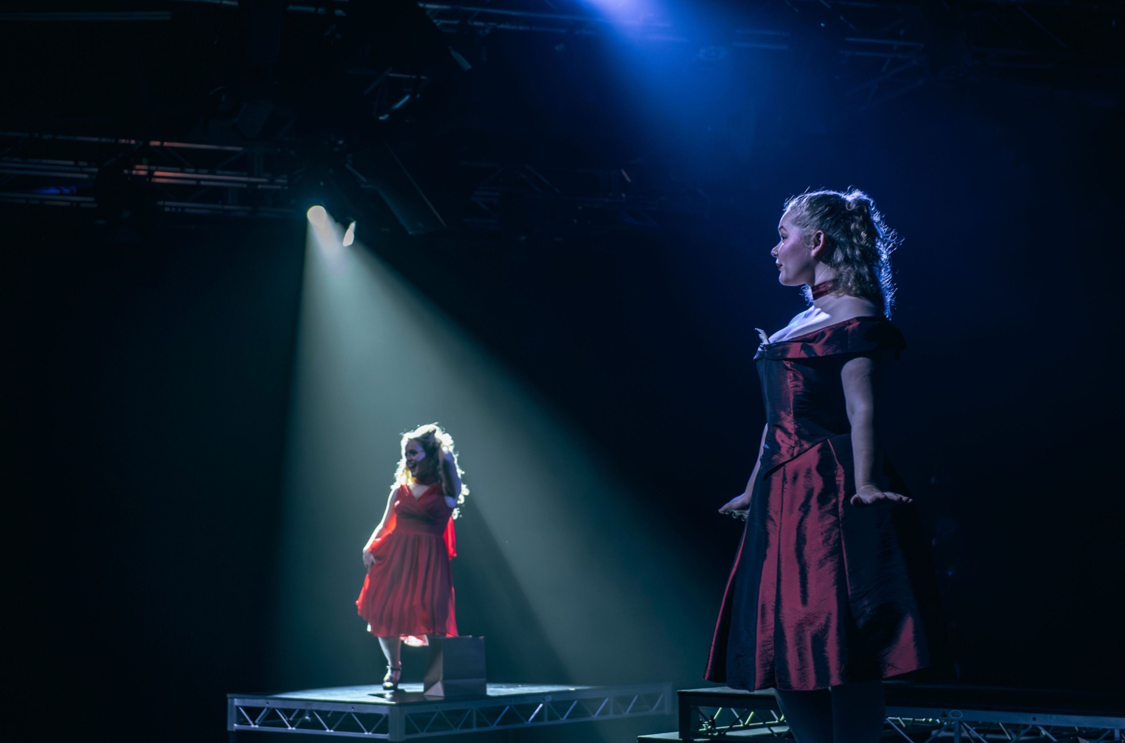 Theatre production photograph showing two actors in red dresses on elevated platforms, lit by spotlight against a dark background.