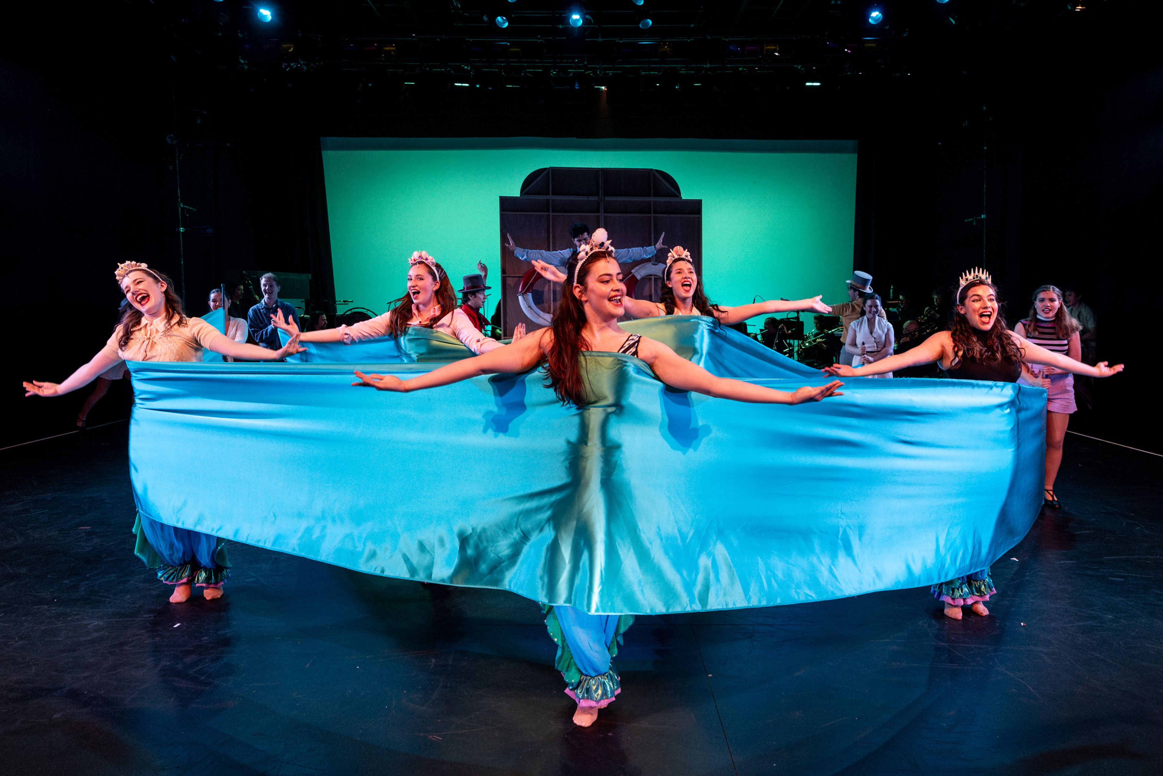 Theatre production photograph showing five performers holding a large piece of turquoise fabric while singing, with dramatic teal lighting and crowns on their heads.