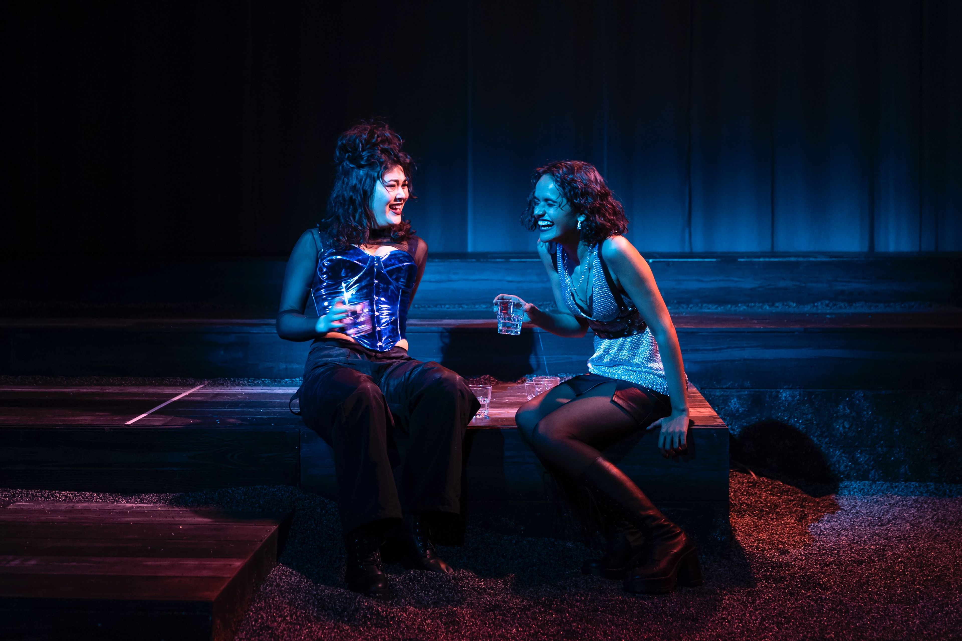 Theatre production photograph of cabaret scene with two performers laughing together on platform steps, one in a metallic corset and another in a sparkly dress, lit with dramatic blue and red lighting.