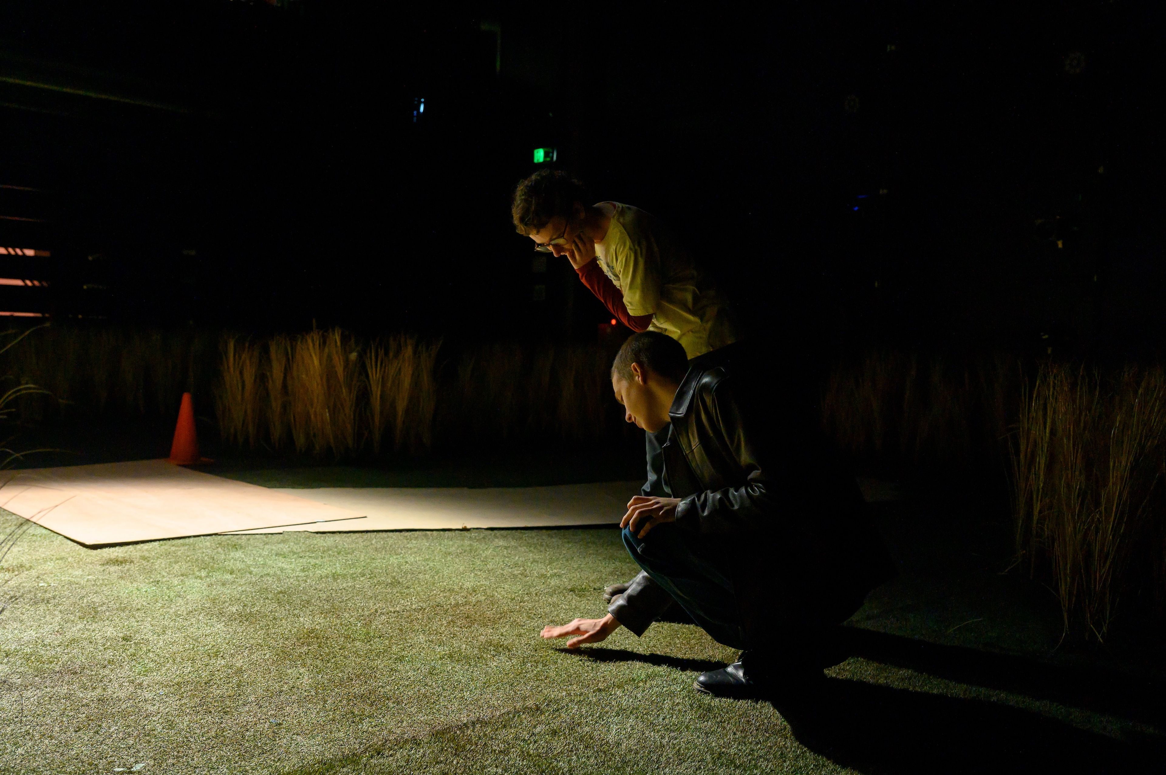 Dimly lit scene showing two crew members on a theatre set with artificial grass, one crouched and inspecting the ground, adding an intimate backstage atmosphere.