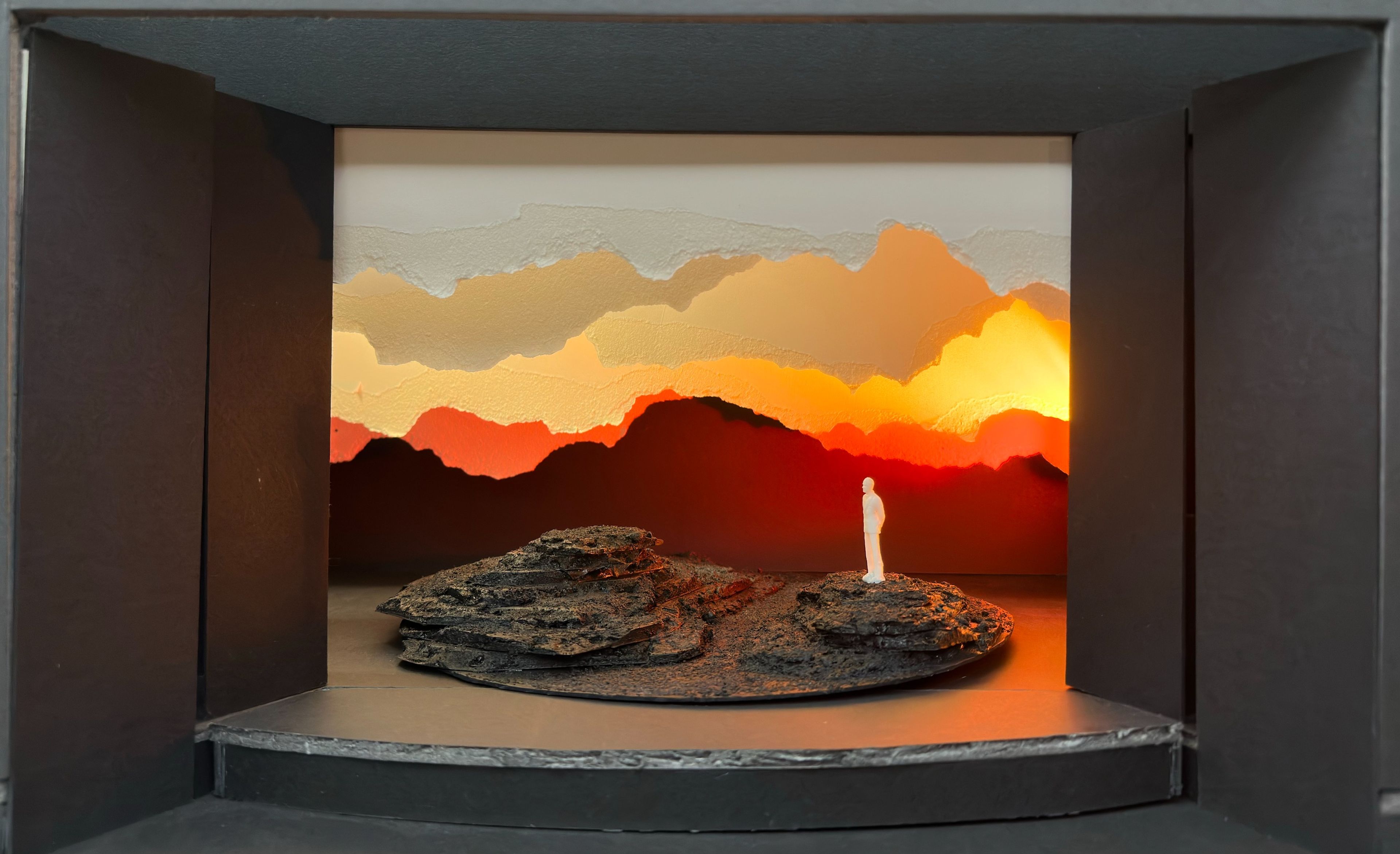 Exhibition setup displaying a scale model set design with paper mountains lit in warm tones.