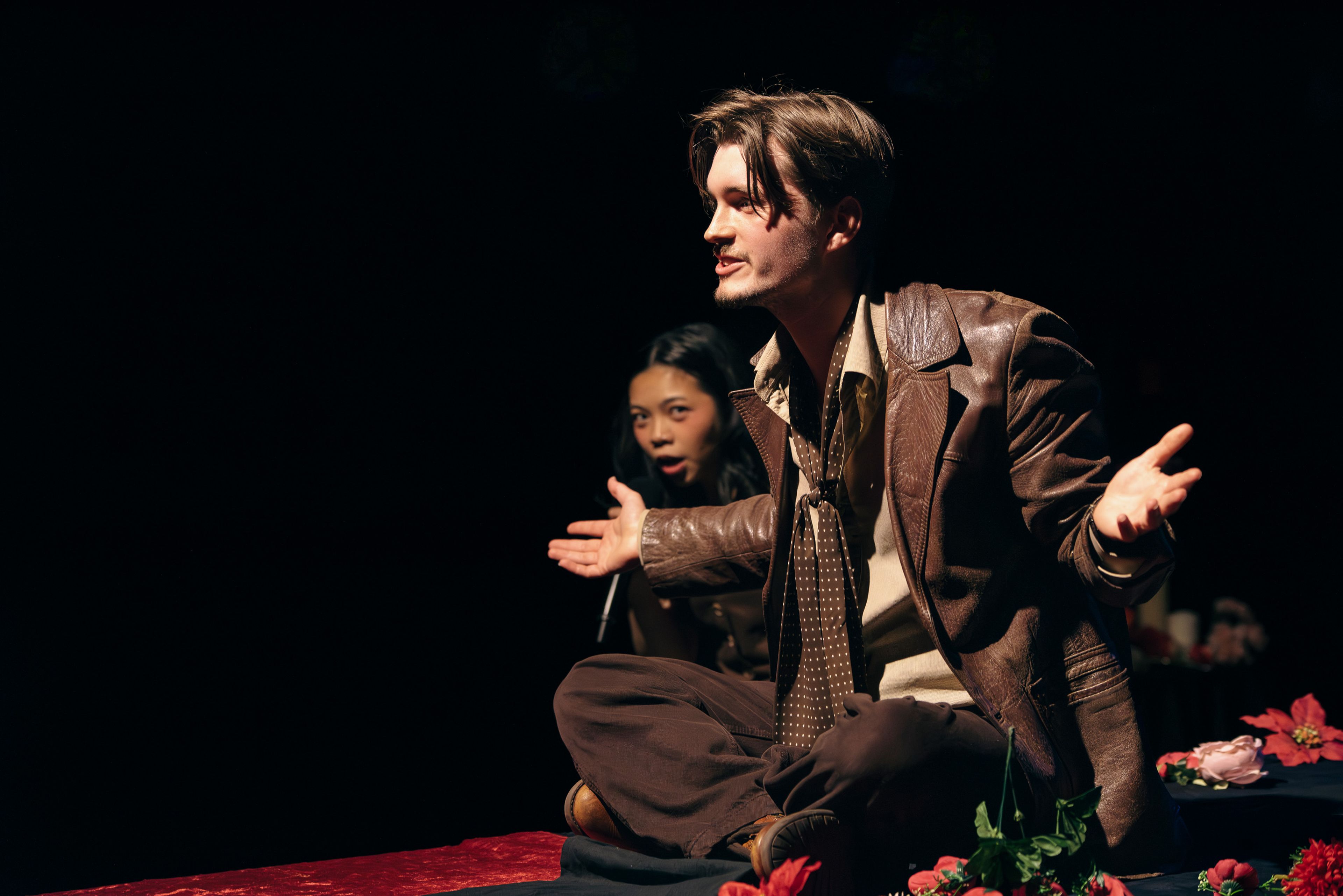 Theatre production photograph showing an actor seated cross-legged on a table, gesturing with open arms, with another actor partially visible behind him.