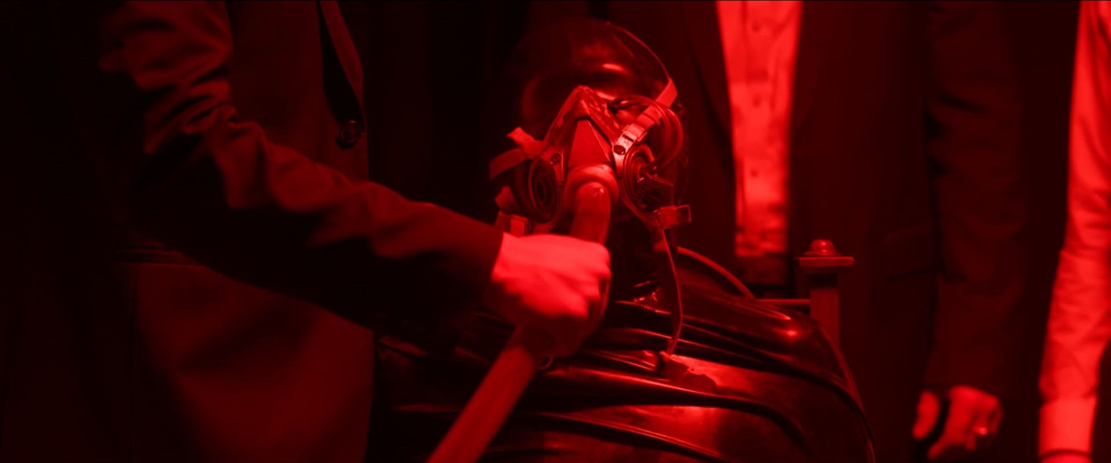 Film still bathed in red lighting, depicting a person in a gas mask being held by others dressed in dark clothing.