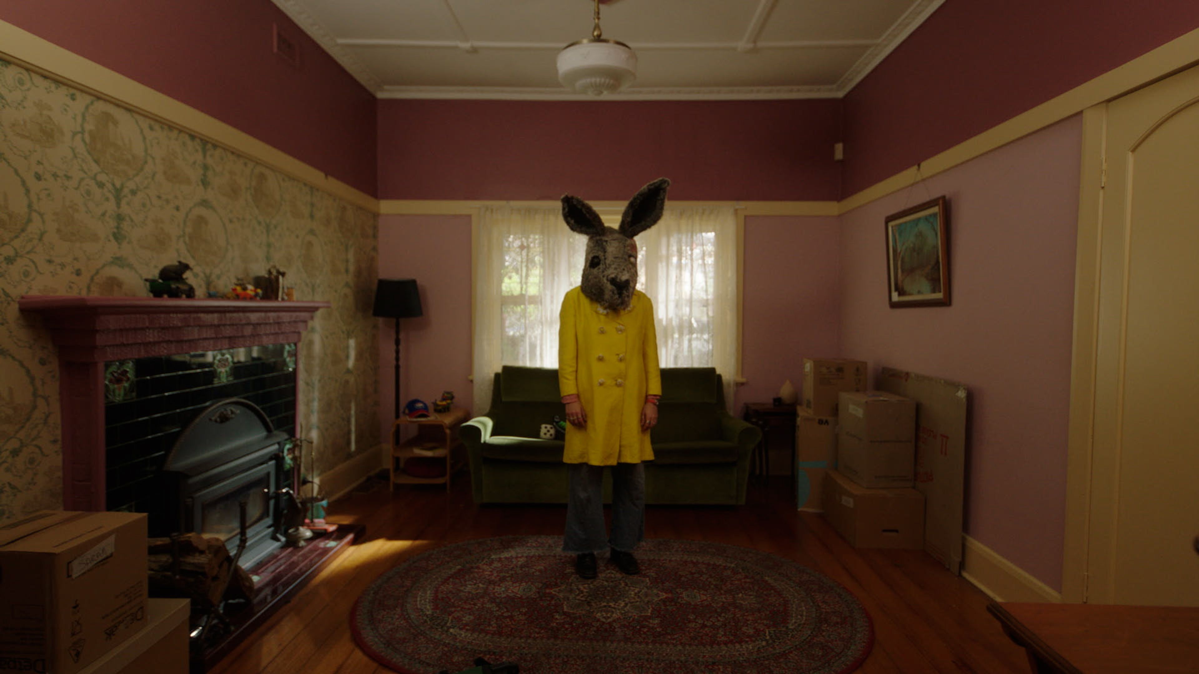 A figure stands wearing a bright yellow coat and a large kangaroo mask in the center of a vintage-style living room with mauve walls and a patterned rug