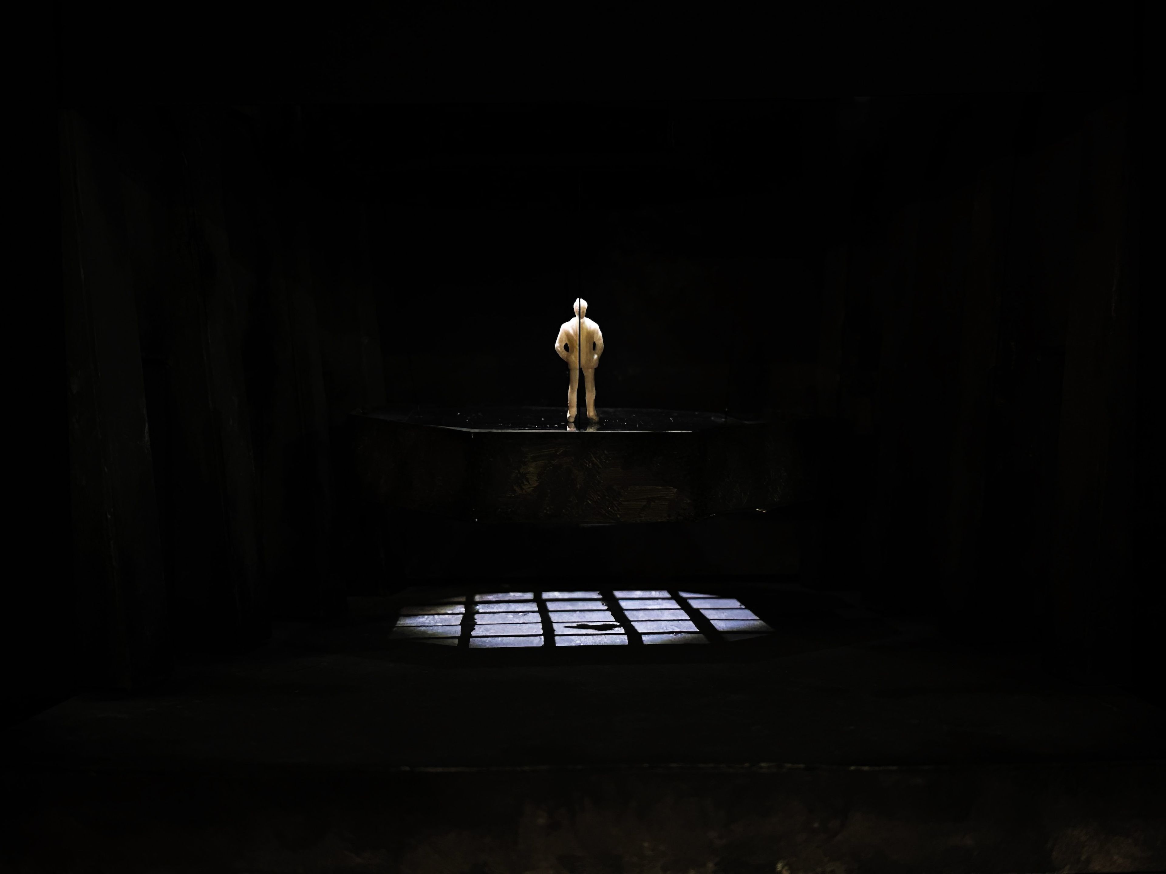 Stage model with a single figure standing on a platform under a spotlight, with a barred window casting shadows on the floor.