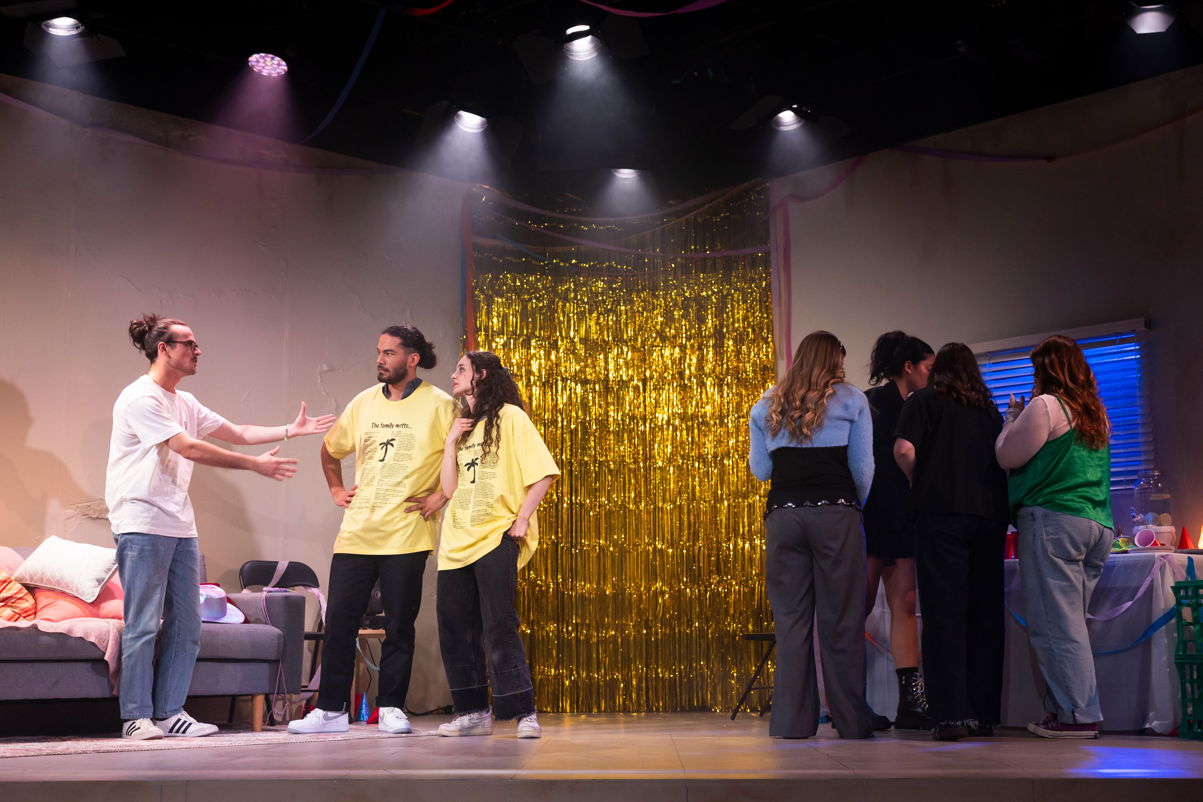 Theatre production photograph in a party setting, with performers in casual clothes having a conversation. Two wear matching yellow t-shirts, and a gold tinsel curtain hangs in the background.