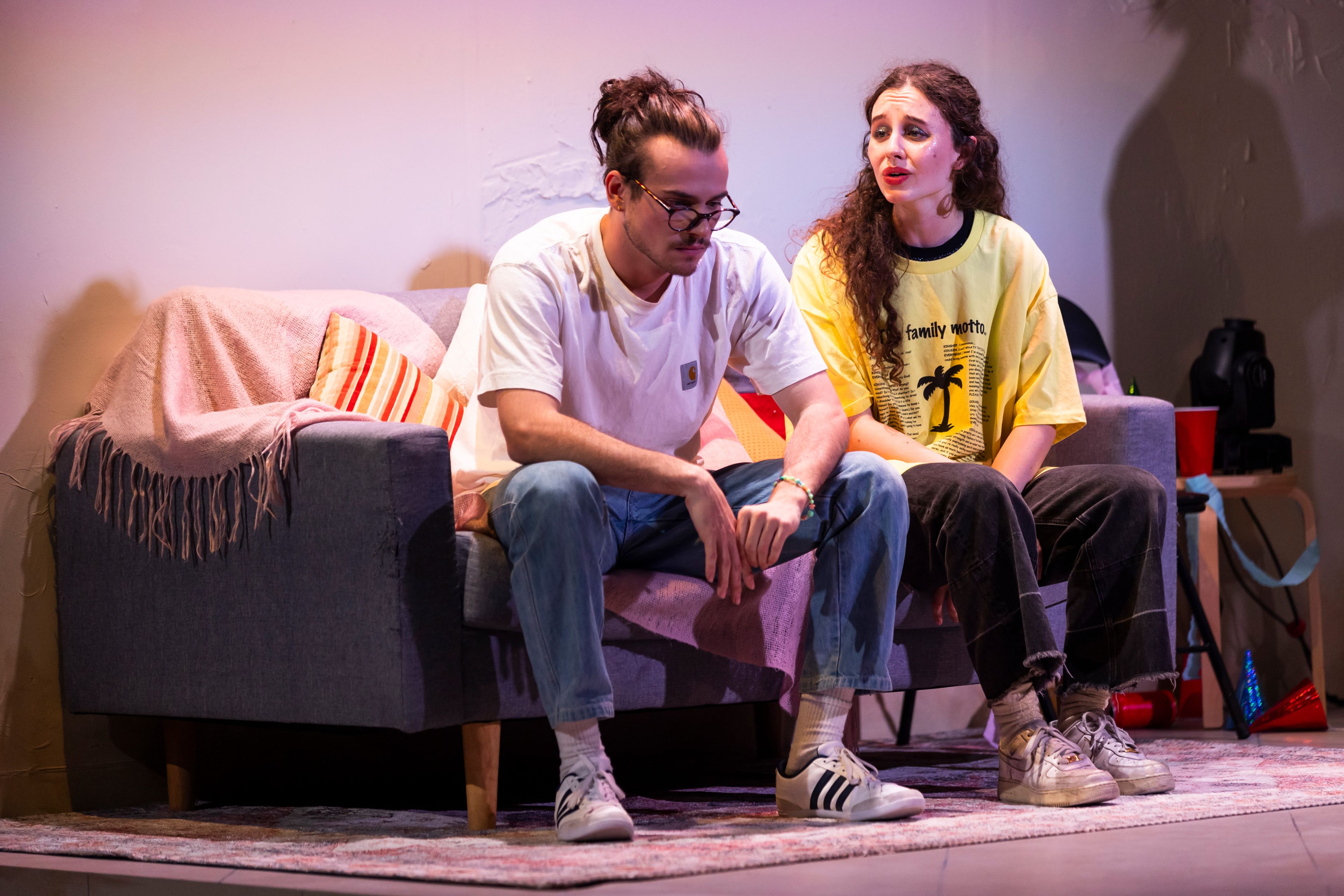 Theatre production photograph of an intimate scene between two performers on a grey couch. One wears a white t-shirt, the other a yellow shirt with a palm tree graphic.