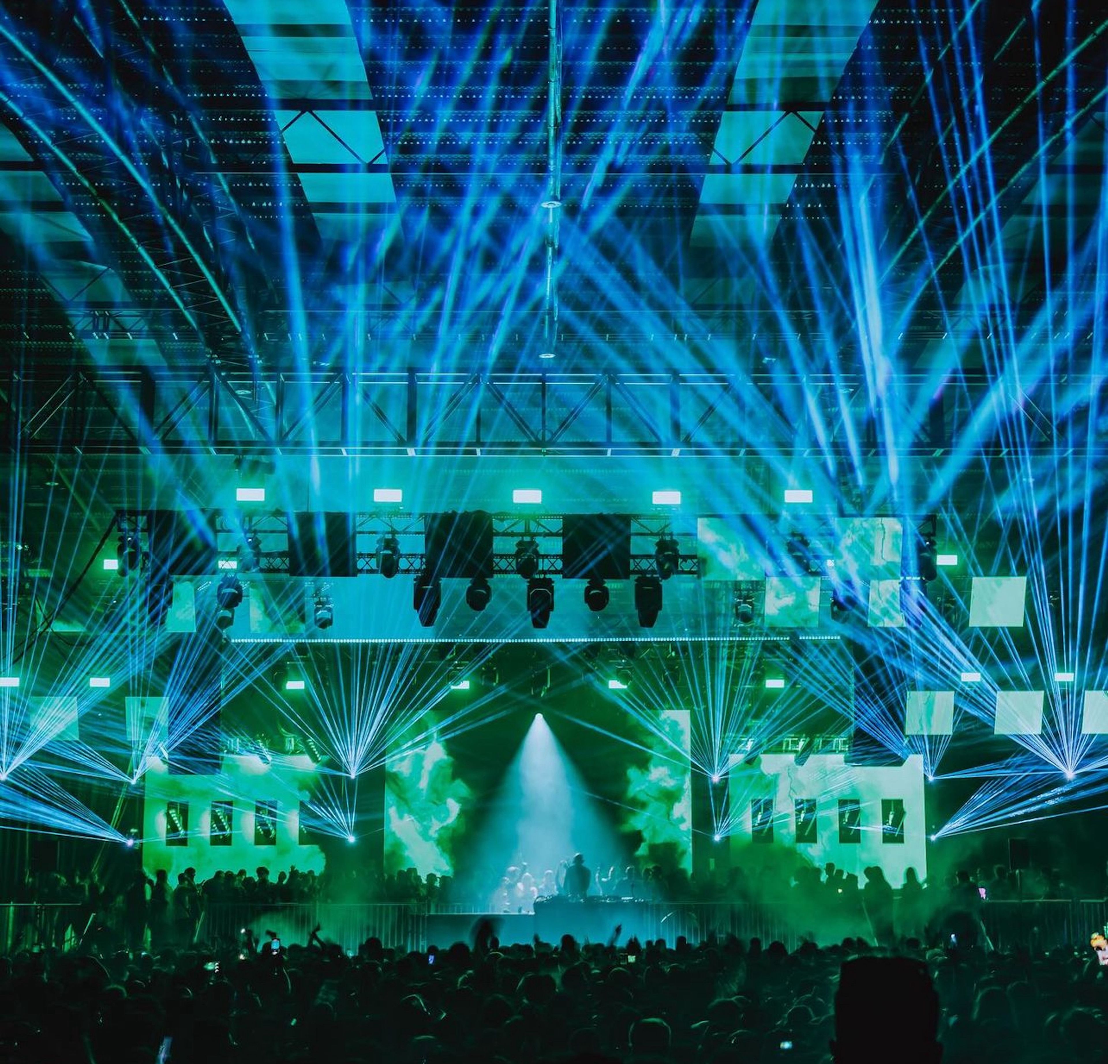 Concert photograph of a stage filled with blue and green laser lights, with an audience silhouetted in the foreground.