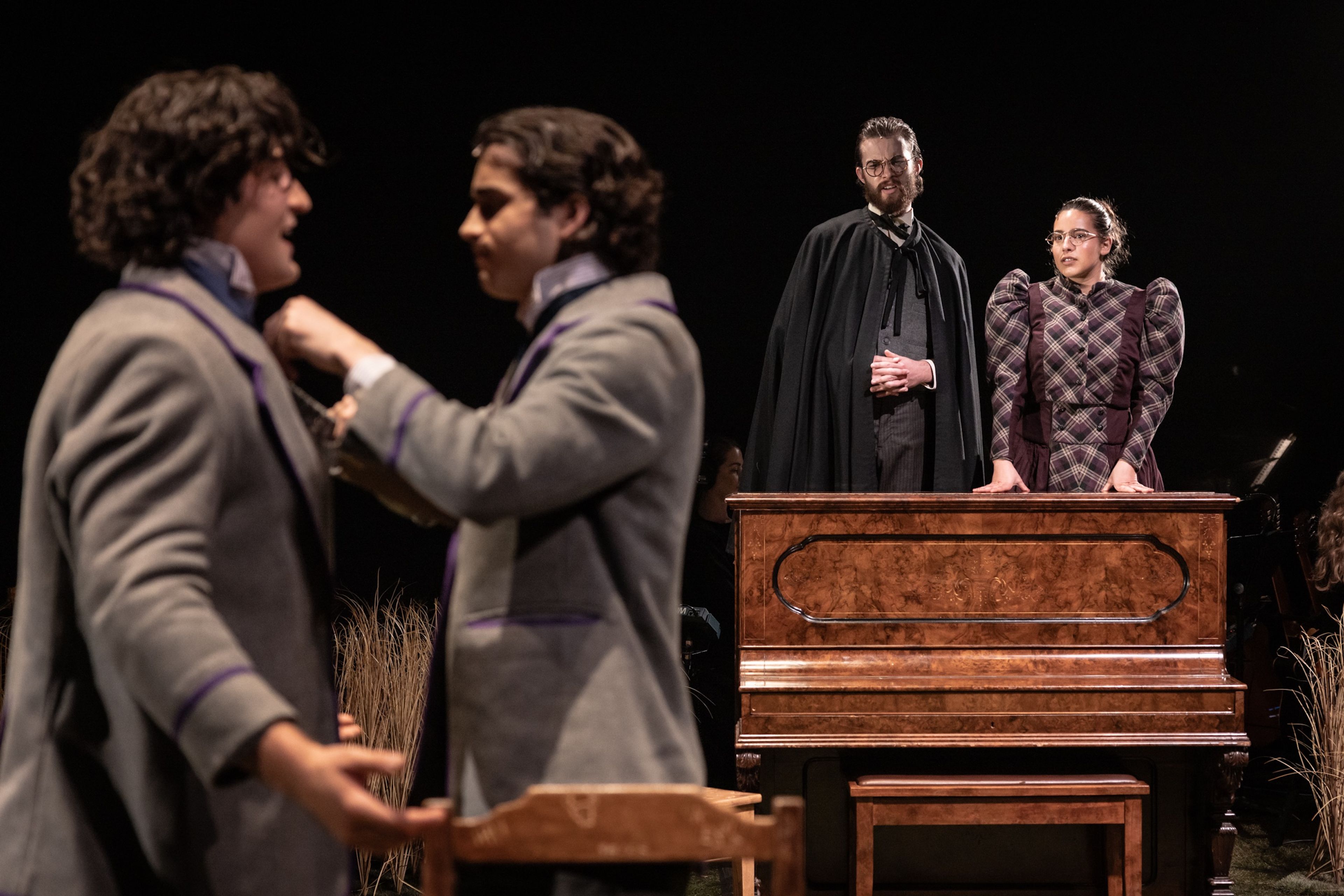 Theatre production photograph showing two students in grey uniforms in foreground, with a figure in a black cape and another in plaid dress standing behind an upright piano.