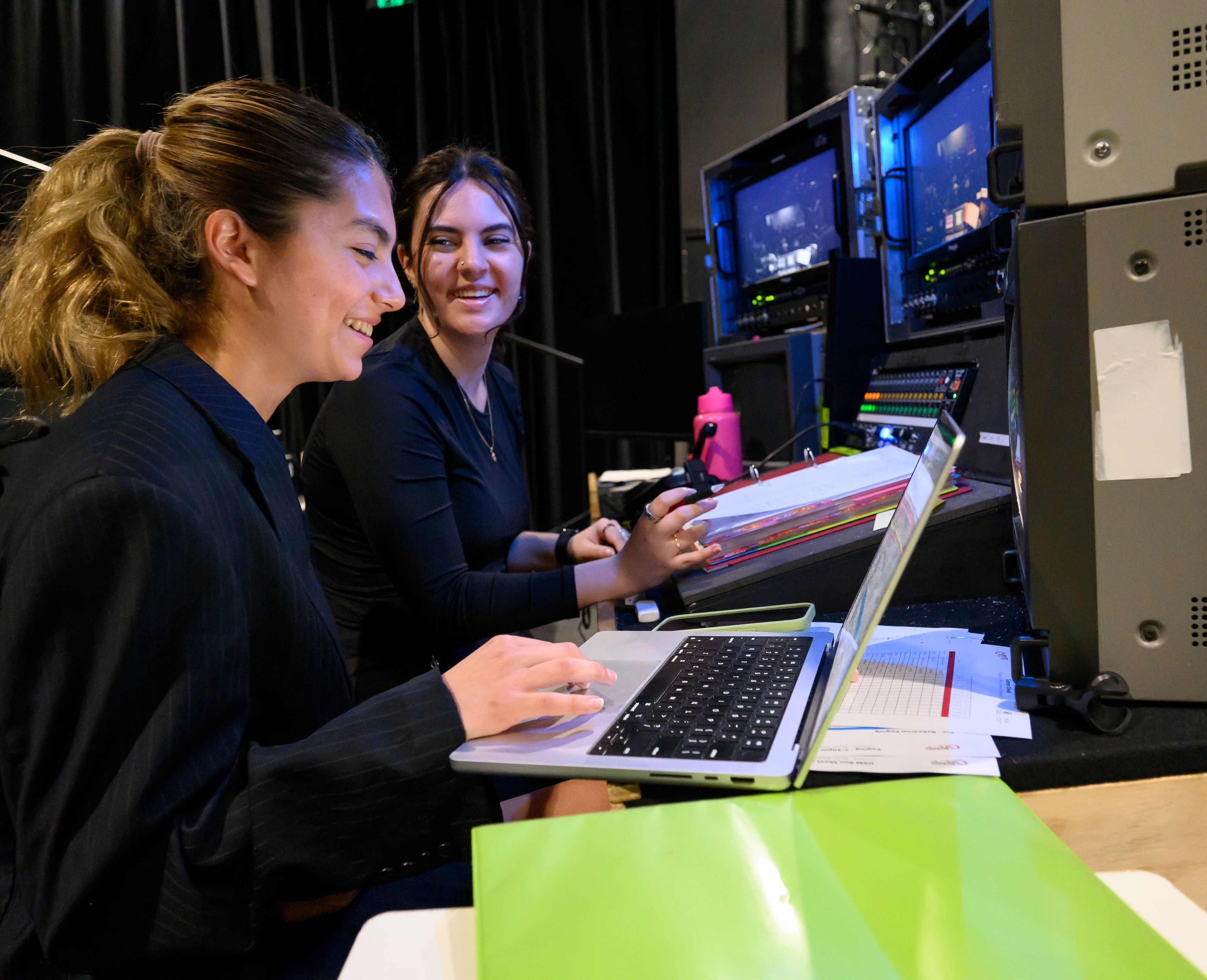 Two crew members smiling and working at a backstage control station with laptops, papers, and screens showing stage activity.