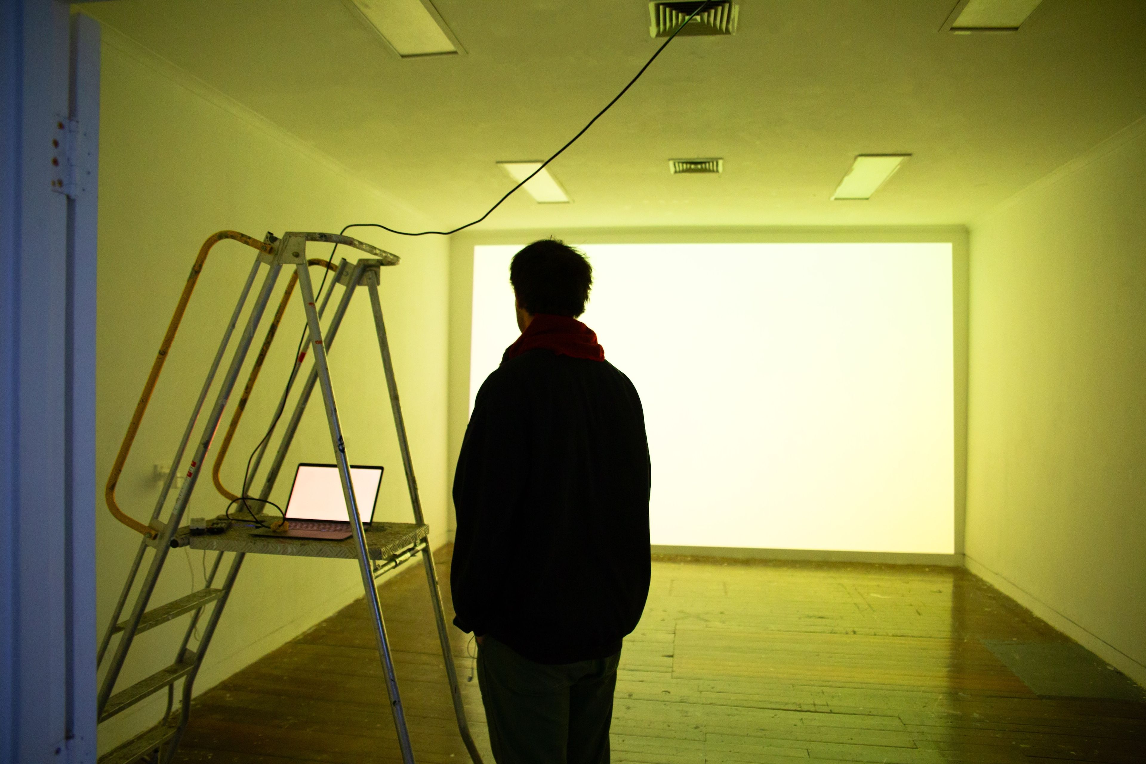 A man stands in a gallery with a yellow-lit projection on the back wall. A ladder with a laptop sits nearby, and a single cord hangs from the ceiling, emphasising a minimalist setup for an immersive visual experience.