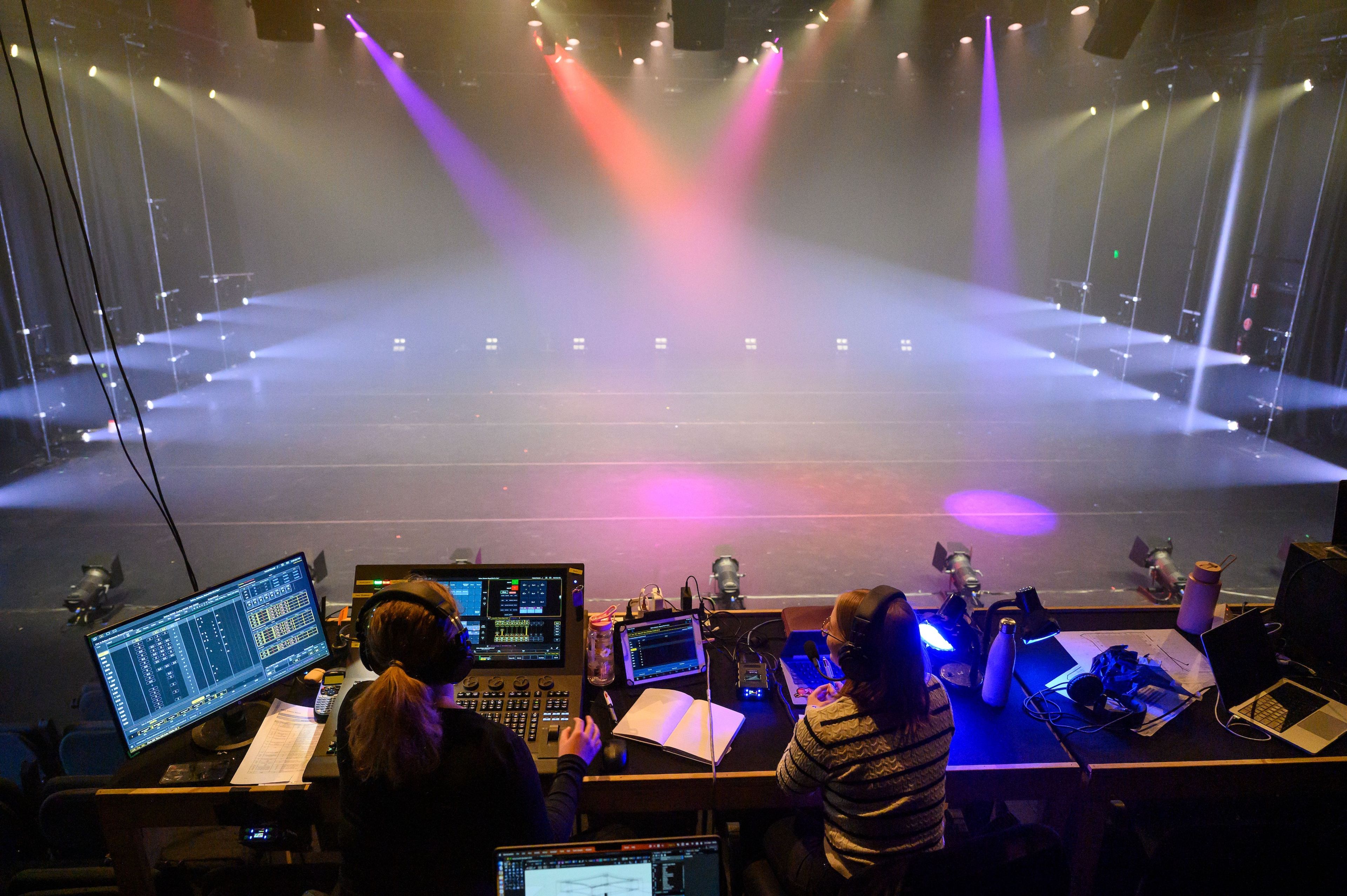 Theatre production photograph showing two crew members seated at a control desk, operating lighting and sound equipment, with a hazy stage lit in pink and purple hues.