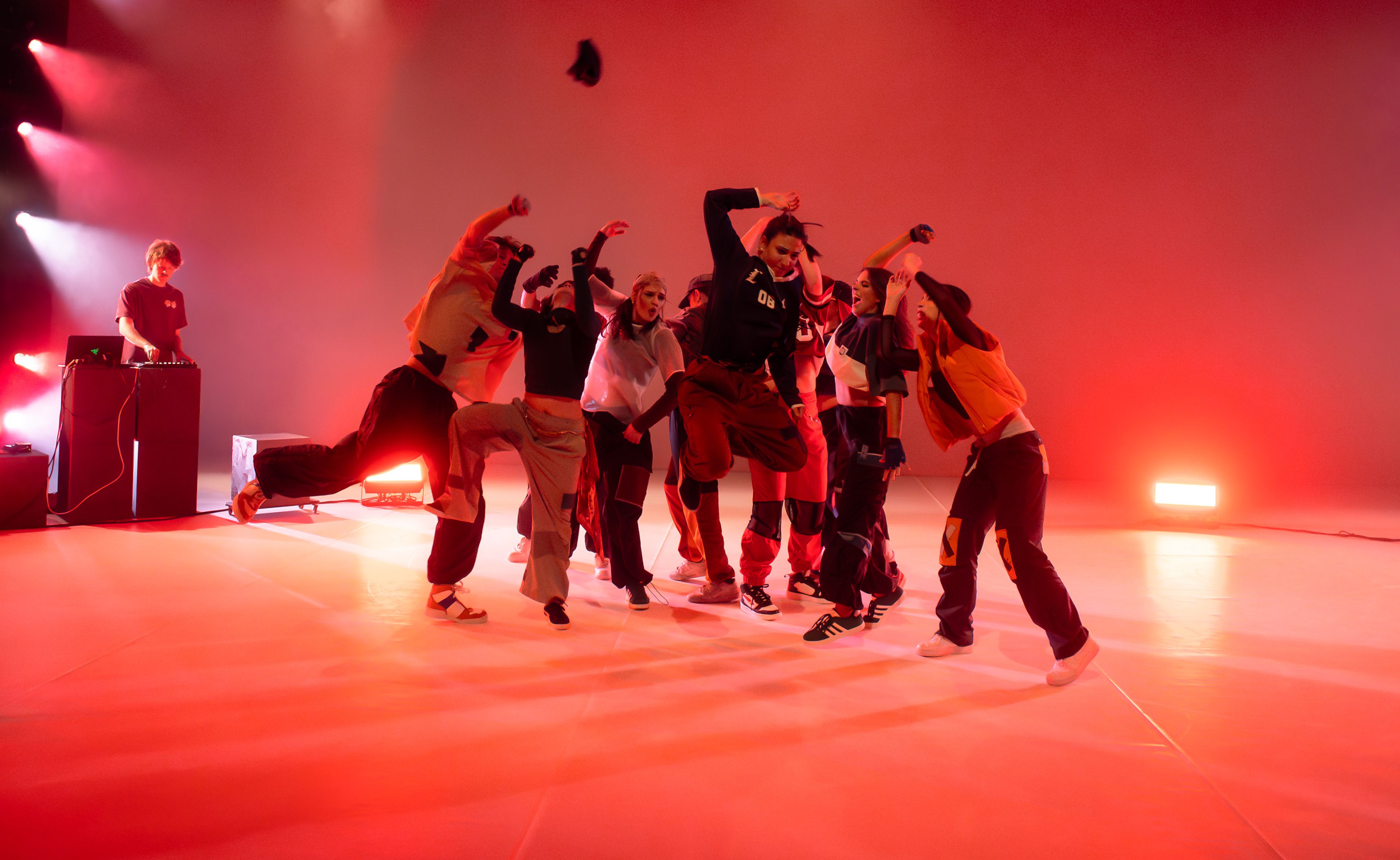 Dance production photograph with a group of dancers mid-motion, lifting their arms and legs in expressive poses, against a warm red-lit background, with a DJ on stage.