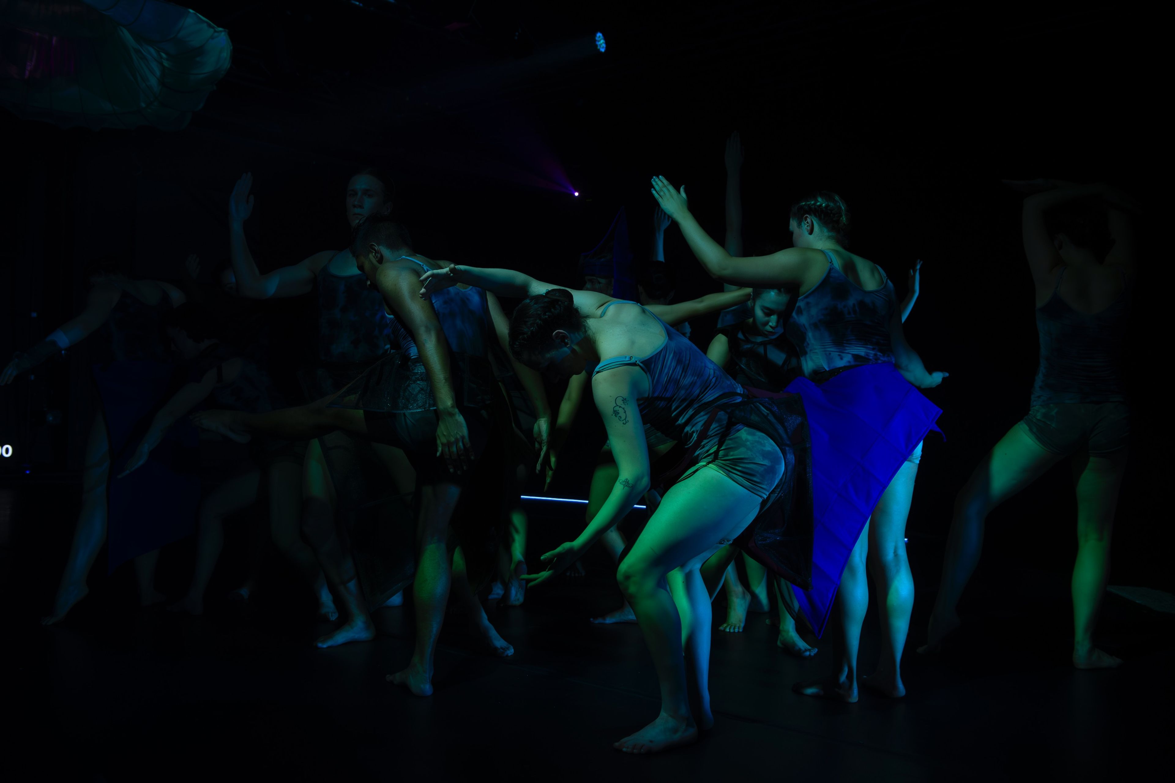 Performance photograph of dancers in blue and green theatrical lighting, creating dynamic movement through bending and reaching gestures