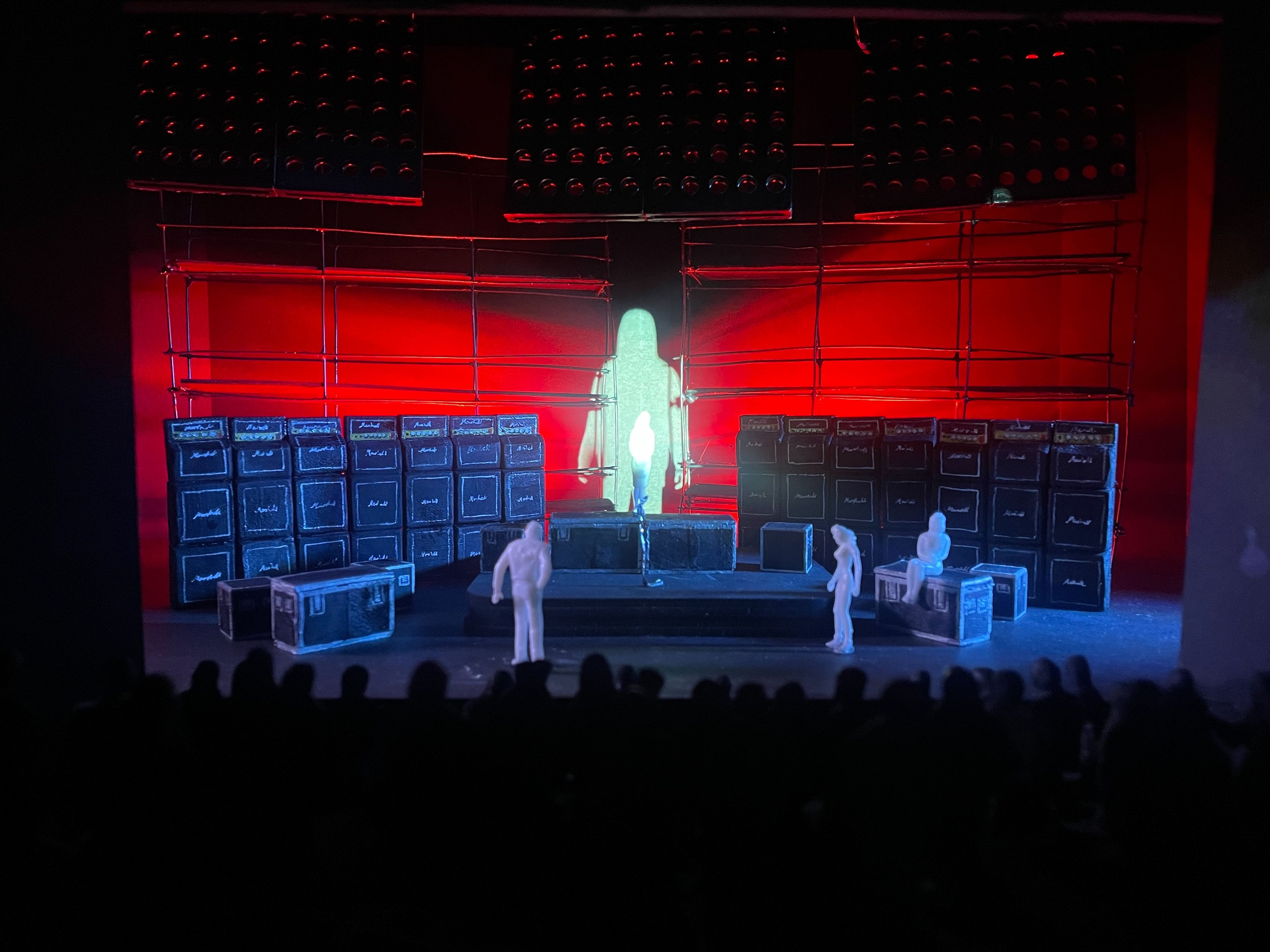 Scale model of a set design featuring a white figure on a stage with a wall of amplifiers and a red-lit backdrop.