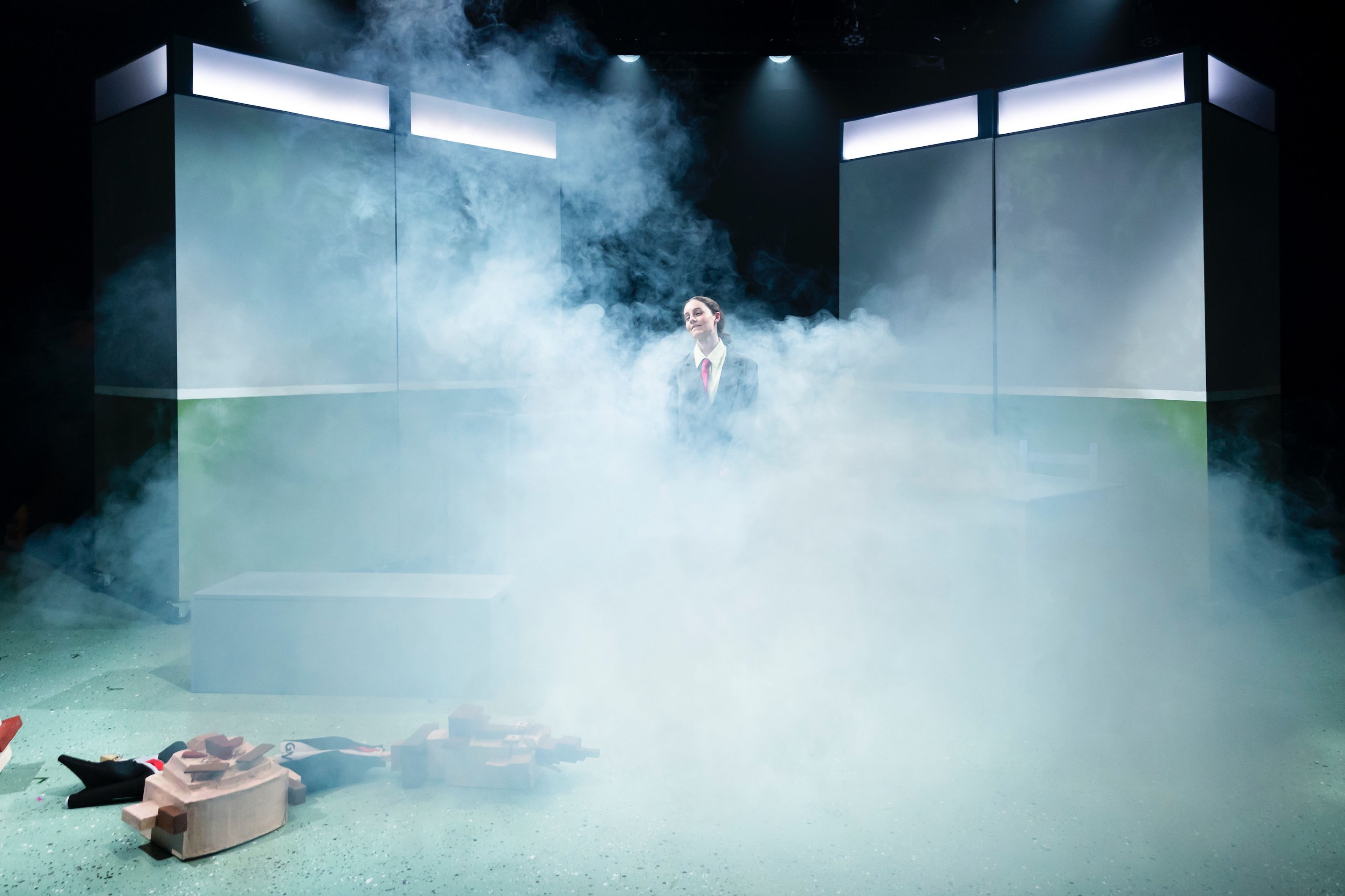 A lone actor stands amidst a cloud of stage fog, surrounded by towering set pieces, under soft overhead lighting.