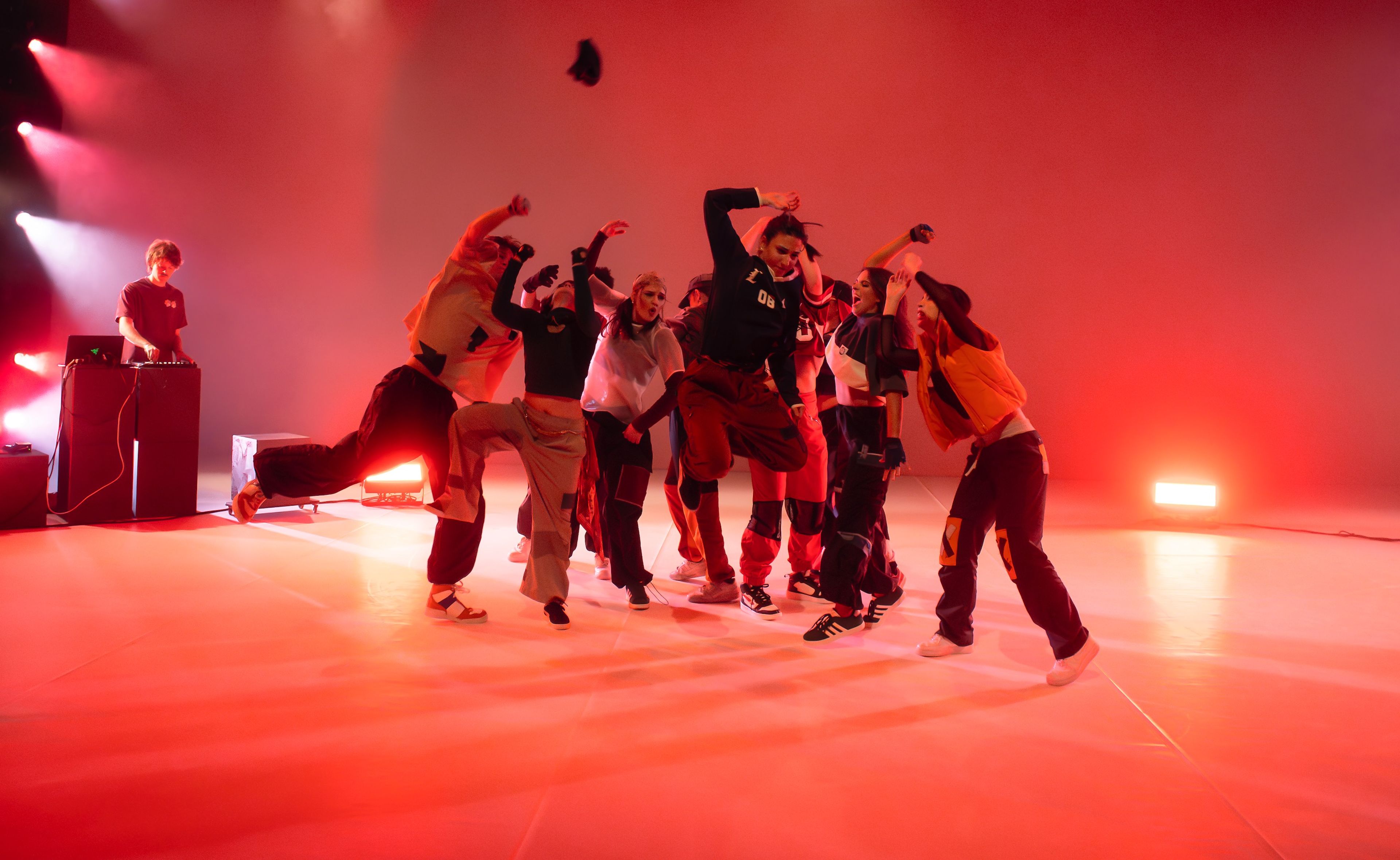 Dance performance photograph of a group of dancers moving dynamically in red lighting, with a DJ set up in the background.