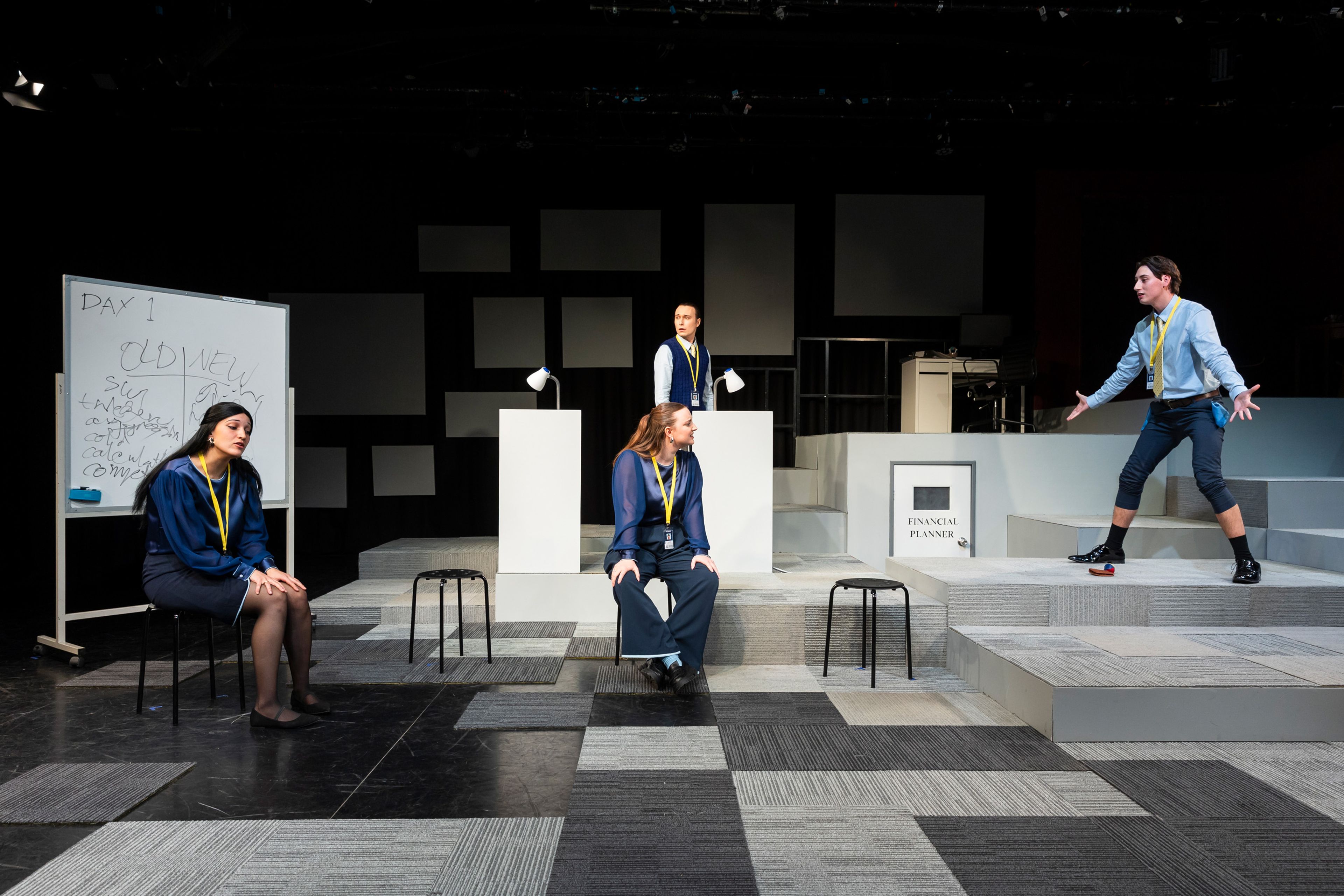 Theatre production photograph featuring four performers in a modern office set with geometric grey carpeting, including a whiteboard marked 'Day 1' and performers in navy blue corporate wear with yellow lanyards.