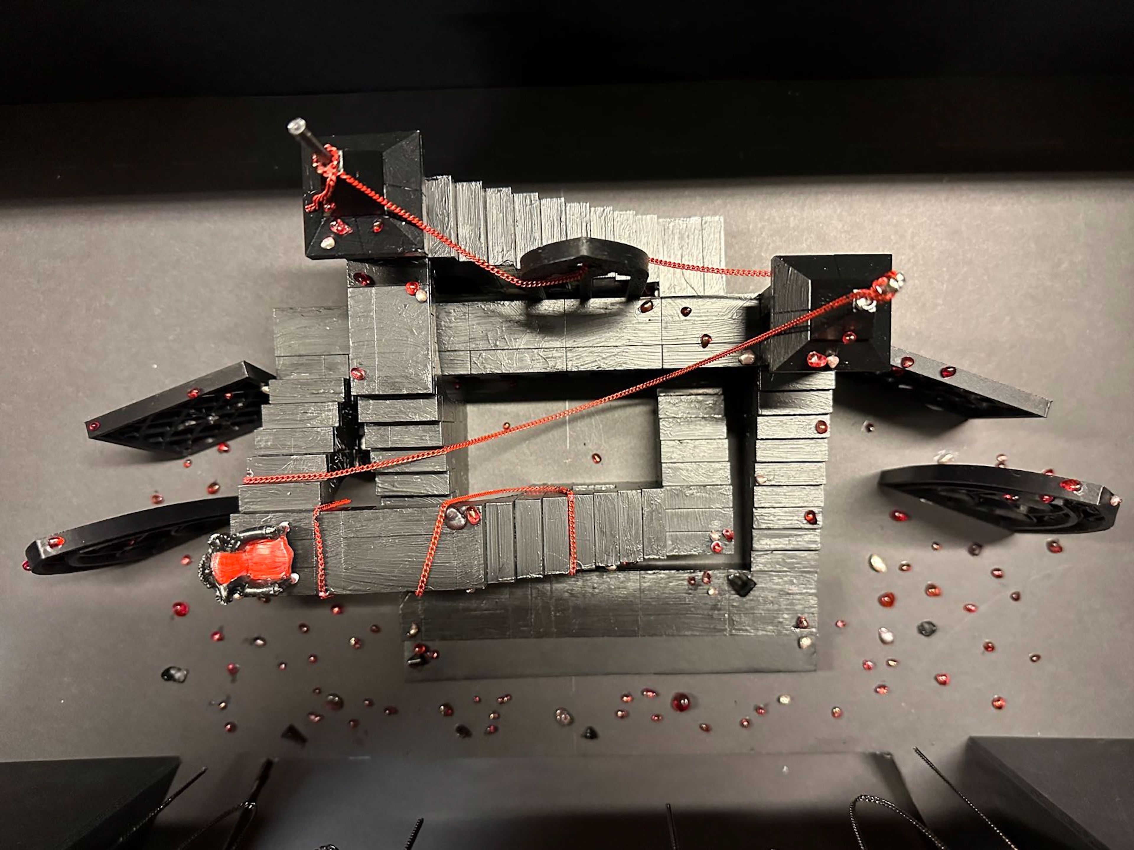 Overhead view of a gothic set design model showing red ropes and scattered red beads on a dark stage.