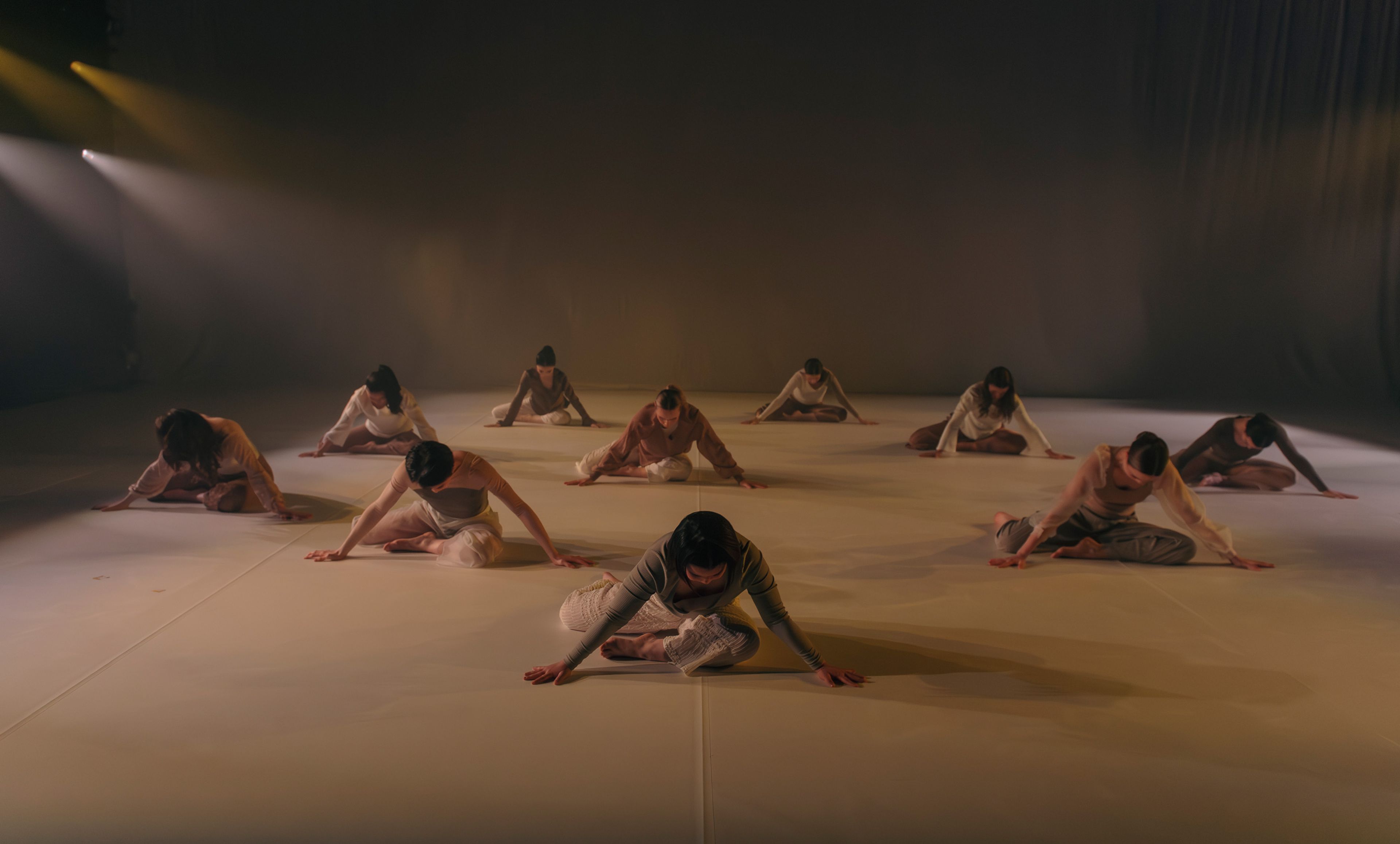 Dance production photograph of multiple dancers sitting on the floor in a spread-out formation, each with a focused, downward gaze, bathed in soft, ambient lighting.