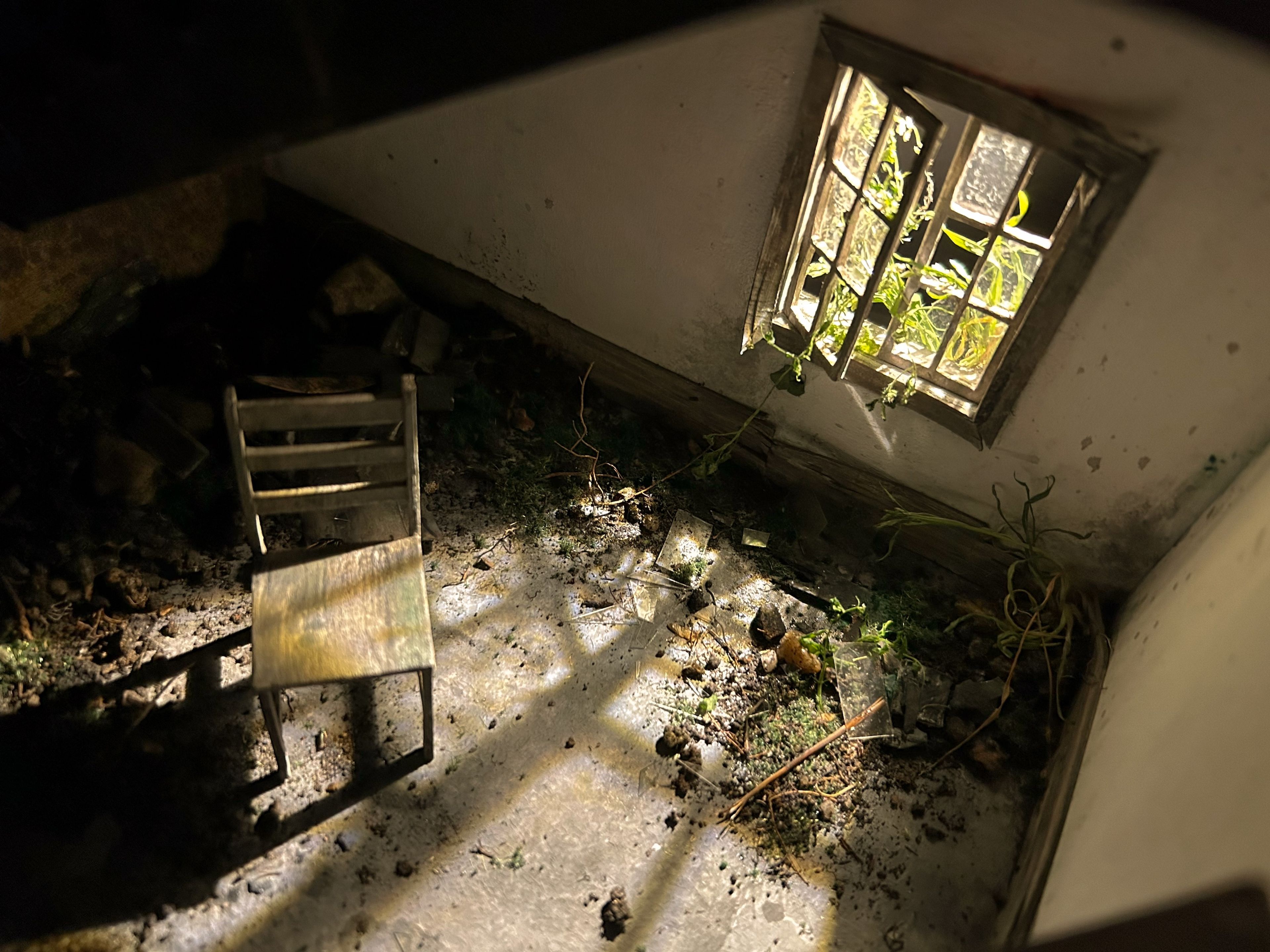 A diorama of a small, abandoned room with a lone wooden chair and a barred window letting in dim light. The room is filled with debris, adding a sense of decay and isolation to the scene.