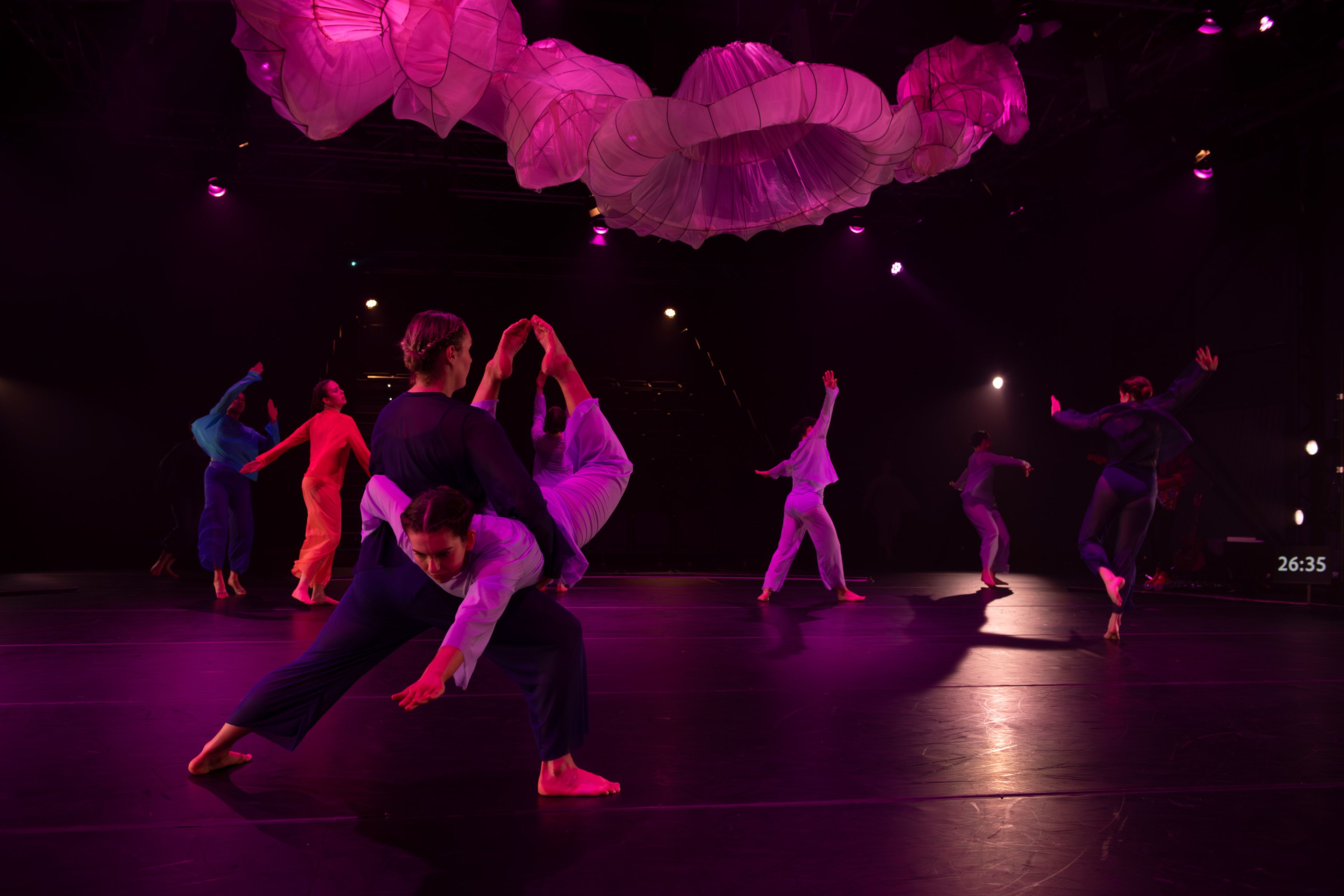 Theatre production photograph featuring dancers in coloured costumes, with one dancer lifted by another, surrounded by suspended pink fabric forms and purple-tinted lighting.