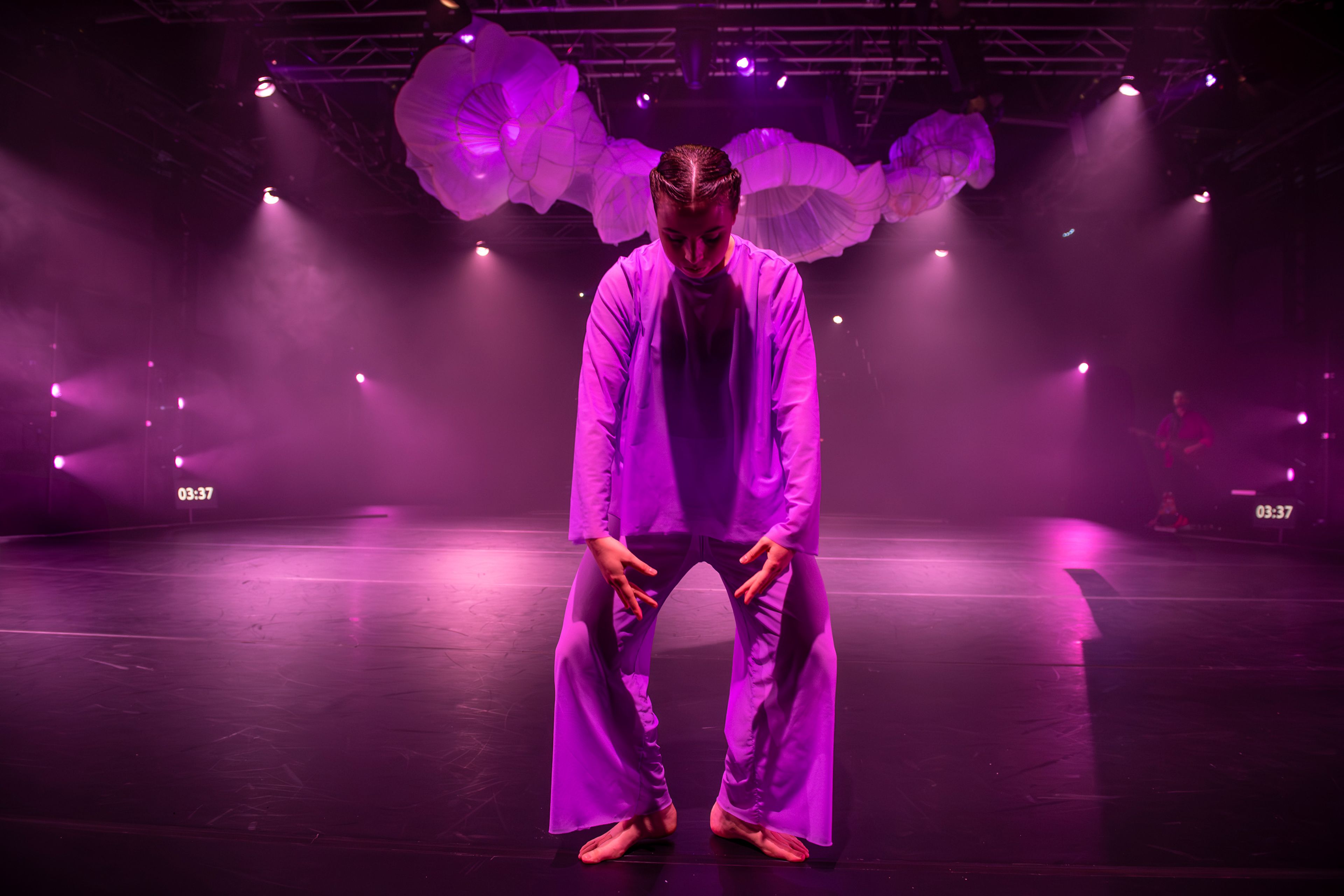 Dance production photograph featuring a solo performer in white costume centred on stage, with sculptural fabric installation overhead, all illuminated in magenta-coloured theatrical lighting with atmospheric haze.